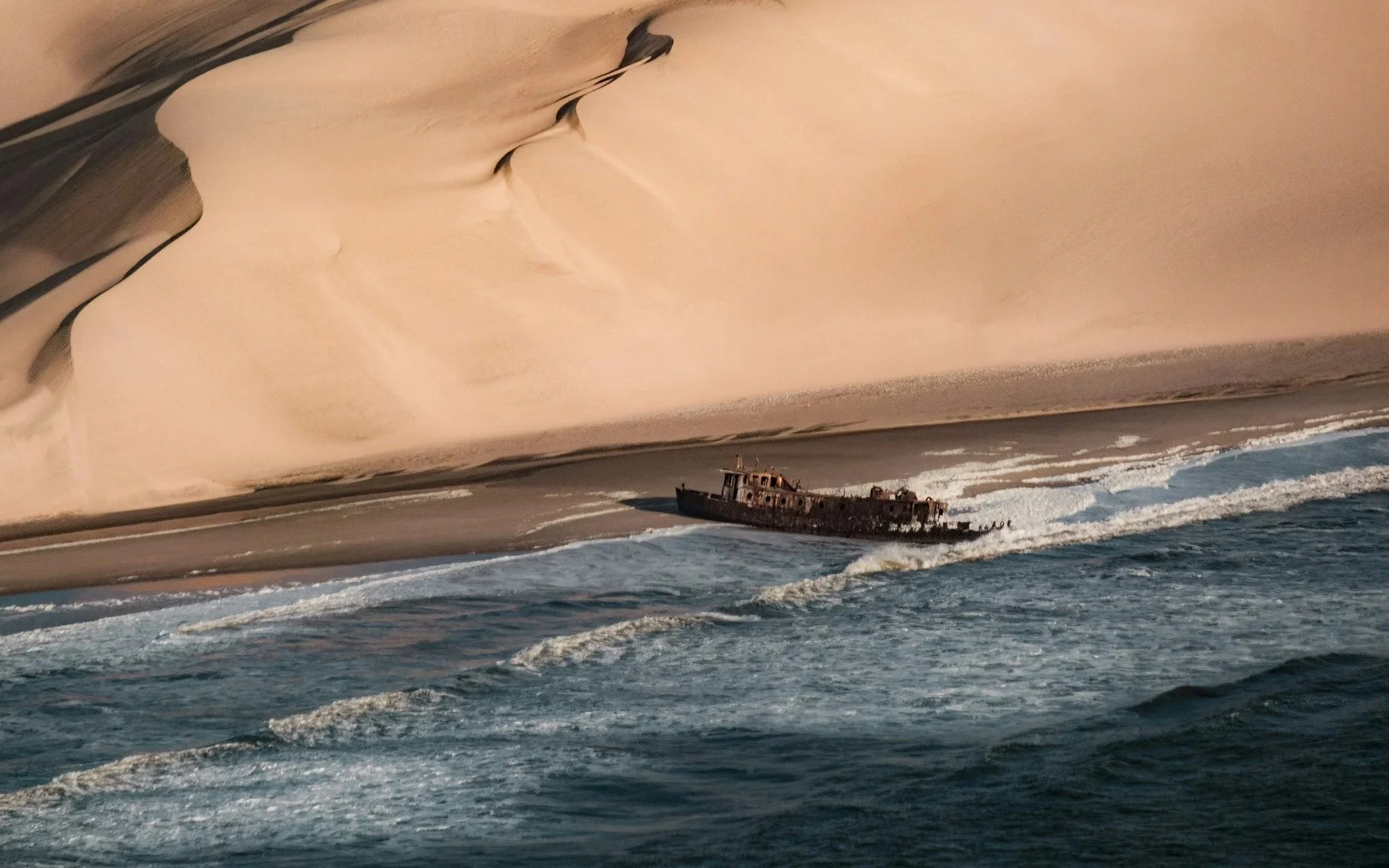 A large shipwreck on a beach with tan sand, with the ship appearing to be old and rusted, partially submerged in shallow ocean waves.