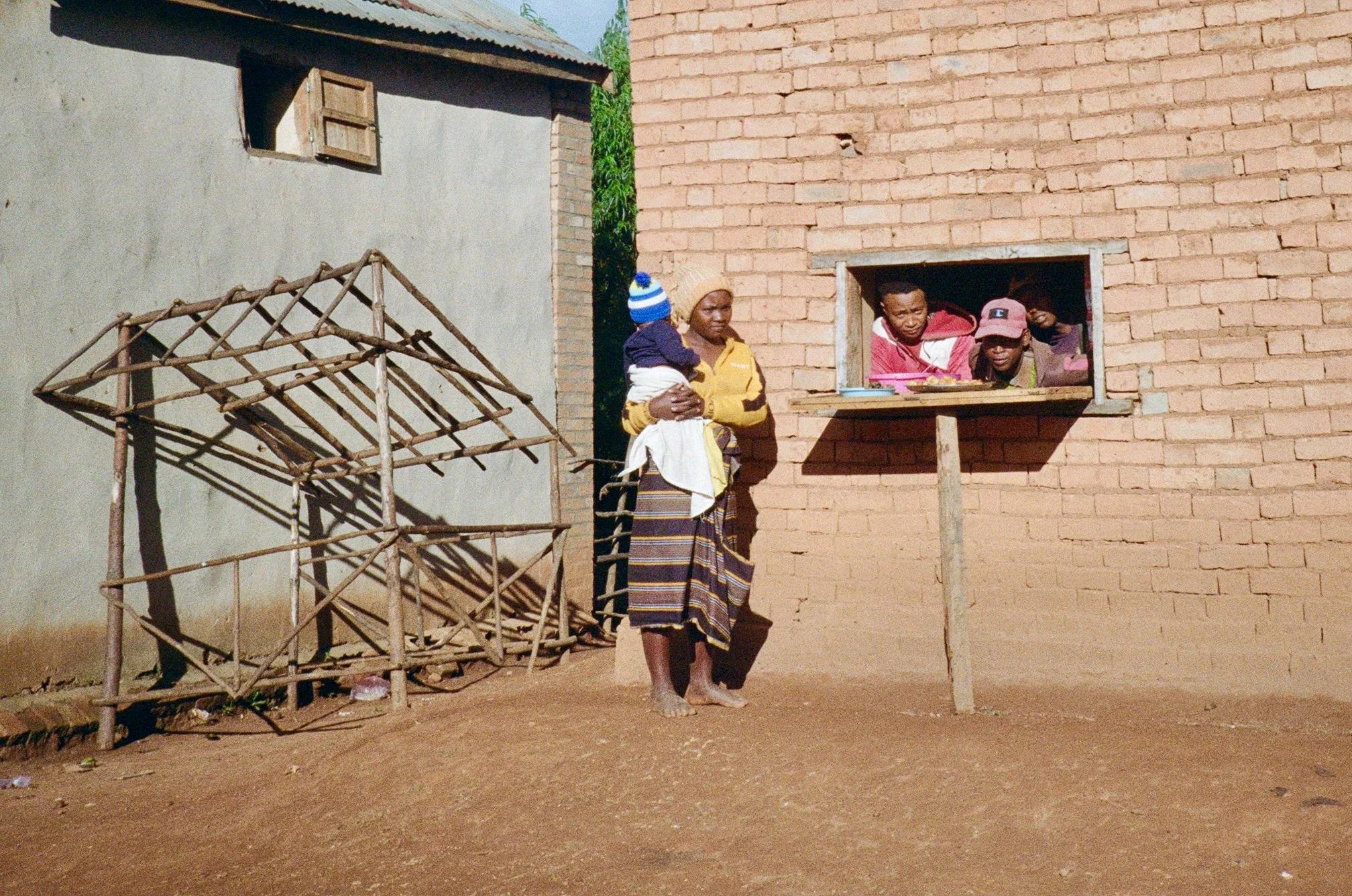 A woman holding a baby standing outside a brick building with a small window, with two people looking out from the window, and a wooden structure on the ground nearby.