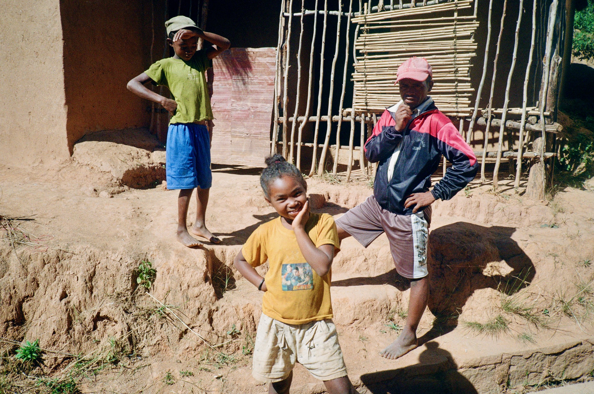 Three children with dark skin and one with light skin, standing outside near a mud house with a bamboo fence, posing and smiling in a sunny environment.