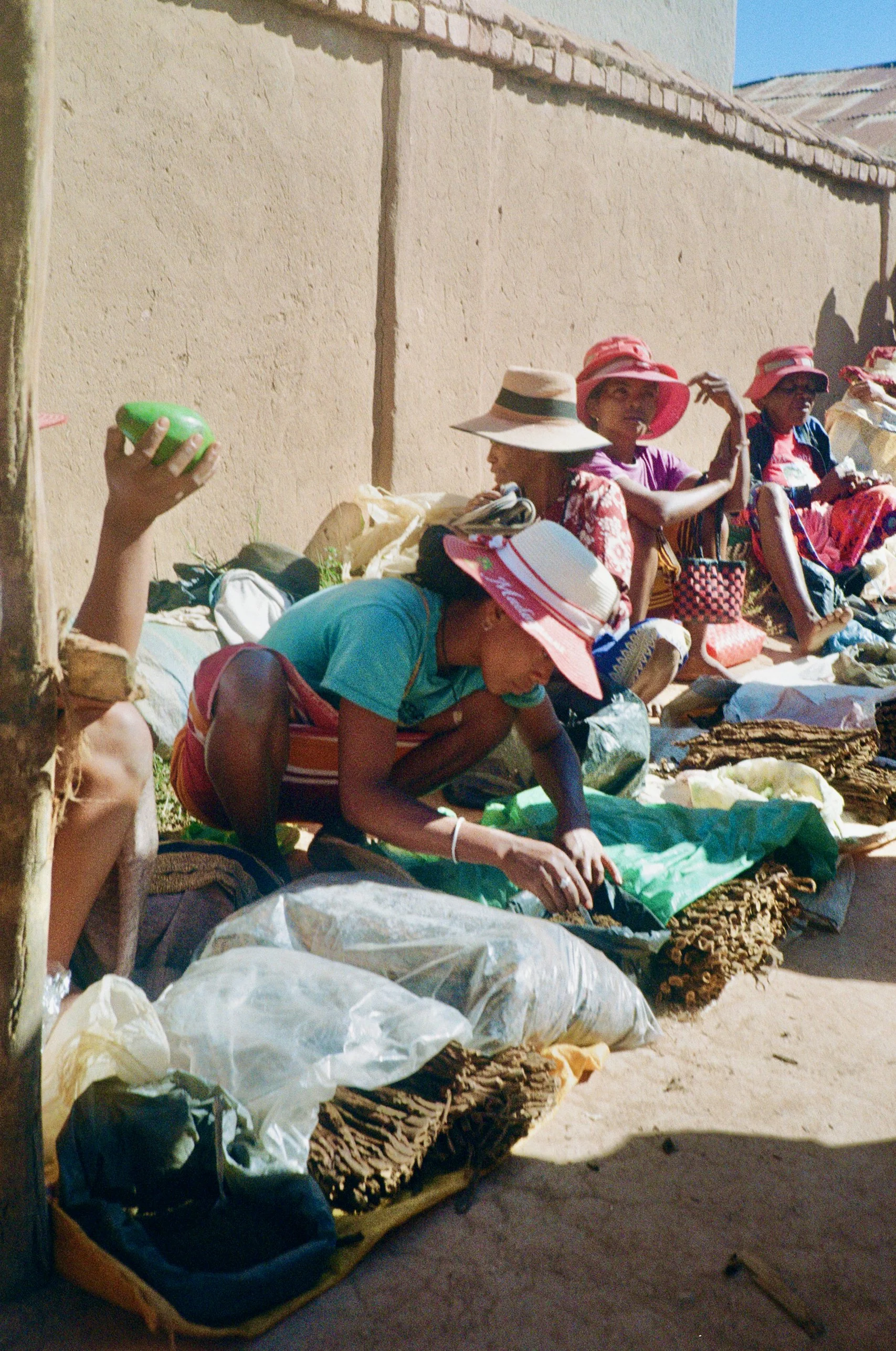 A group of women sitting on the ground against a beige wall, wearing hats, with bags and supplies around them in an outdoor setting.