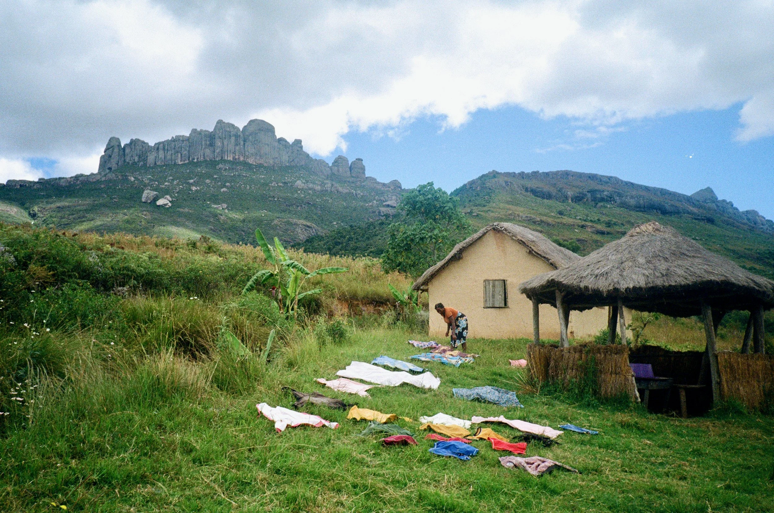 A woman hanging laundry outside a small house with a thatched roof, surrounded by green grass and trees, with a mountain in the background under a partly cloudy sky.