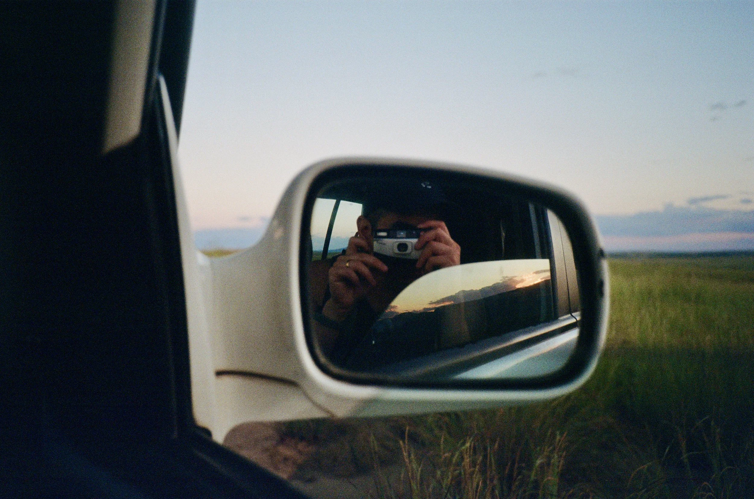 A person's reflection taking a photograph with a camera in the side mirror of a vehicle, with a sunset sky in the background.
