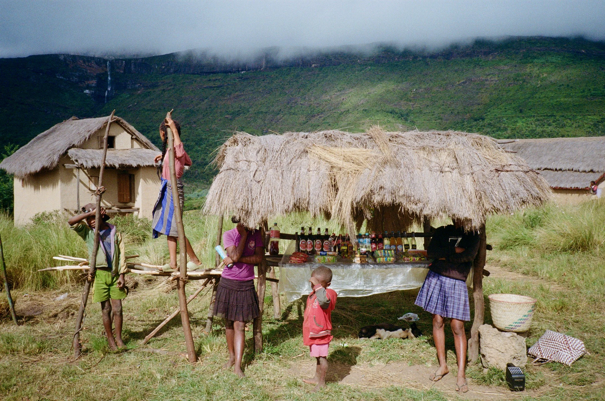 A rural village scene with thatched huts and a small market stall selling snacks and beverages. Several children and a woman are gathered around, some standing and others sitting. Green hills and mountains are visible in the background, partially cov