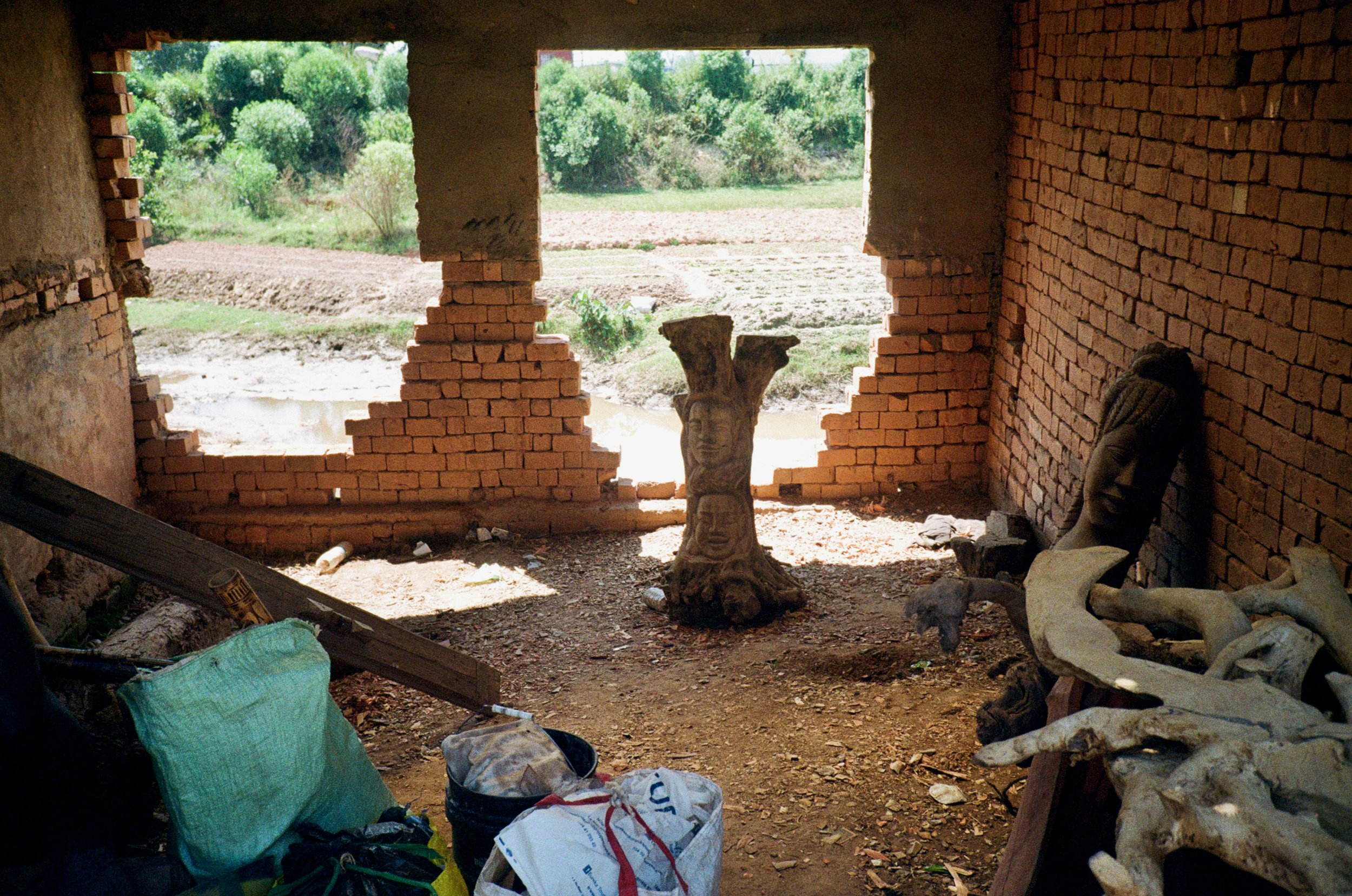 Inside a rustic brick structure with large openings overlooking a green, rural landscape. The interior contains various decorative items, including a carved wooden sculpture of a face and a Buddha head, along with scattered debris and bags on the dir