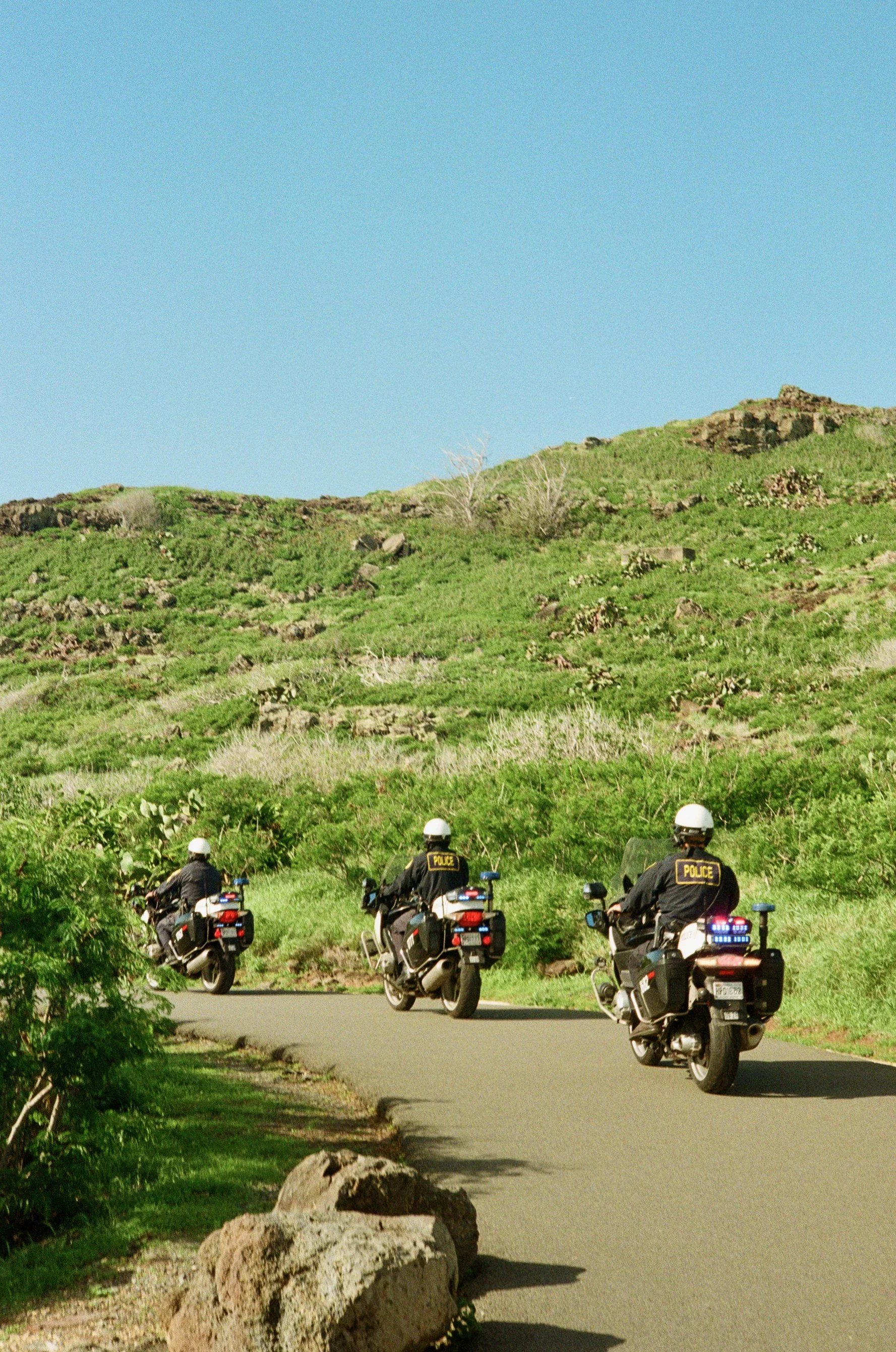Four police officers on motorcycles parked on the side of a curved road, with a green hillside and blue sky in the background.