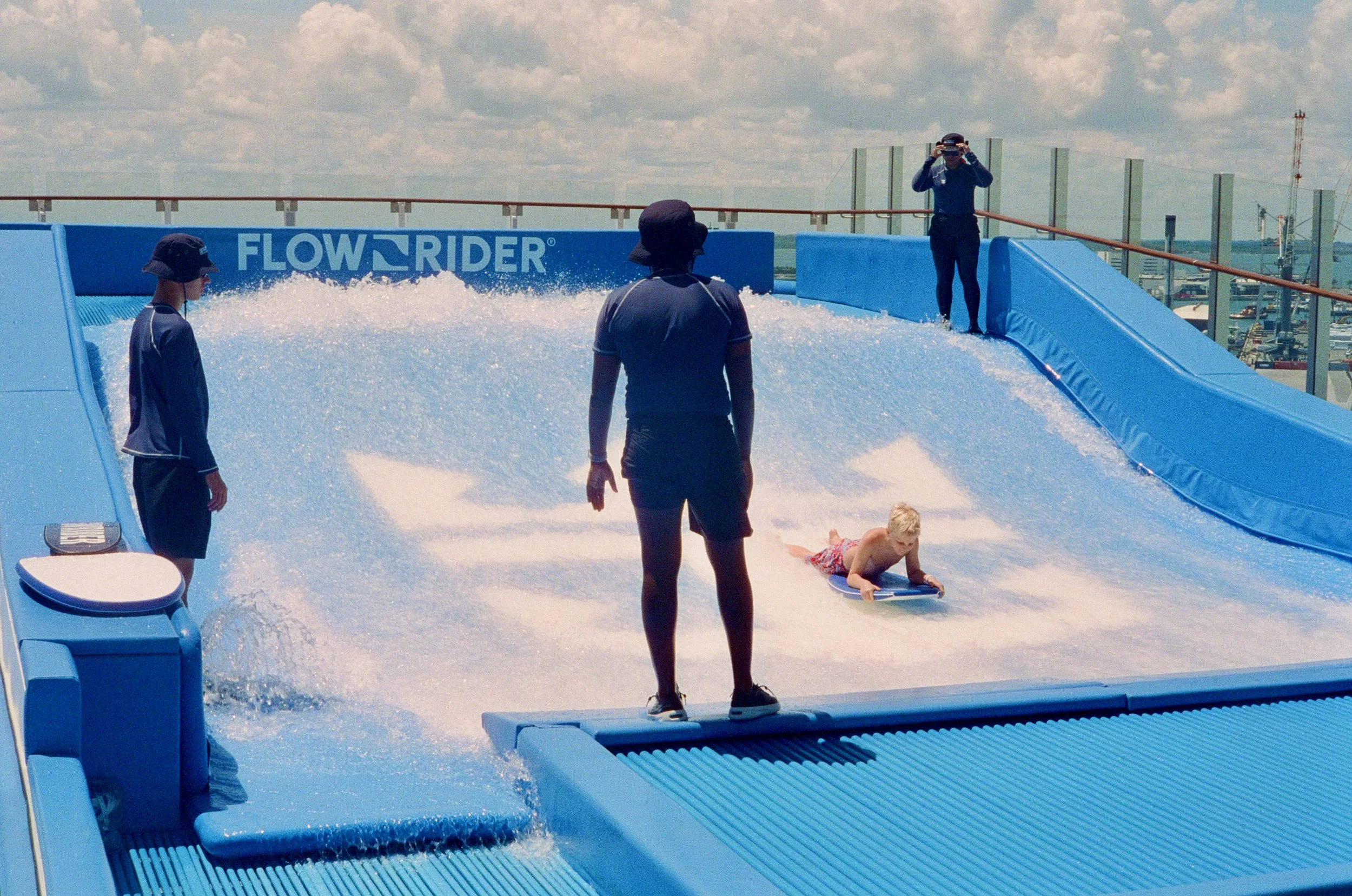 People at a wave pool with a large artificial wave set, children and adults enjoying the water, and a person taking photos, with a city skyline in the background.