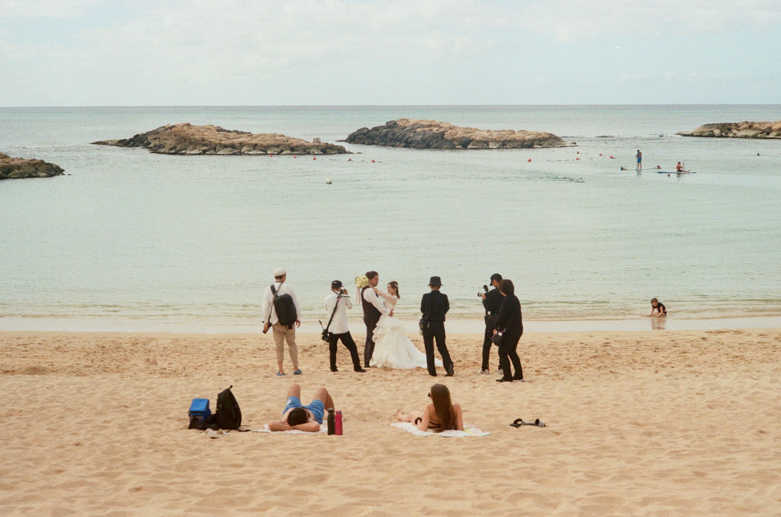 People on a sandy beach near the water with a rocky island and several swimmers in the background. A group of people are gathered, with some sitting on the sand, some standing, and a couple in wedding attire. Others are walking or sitting by the wate