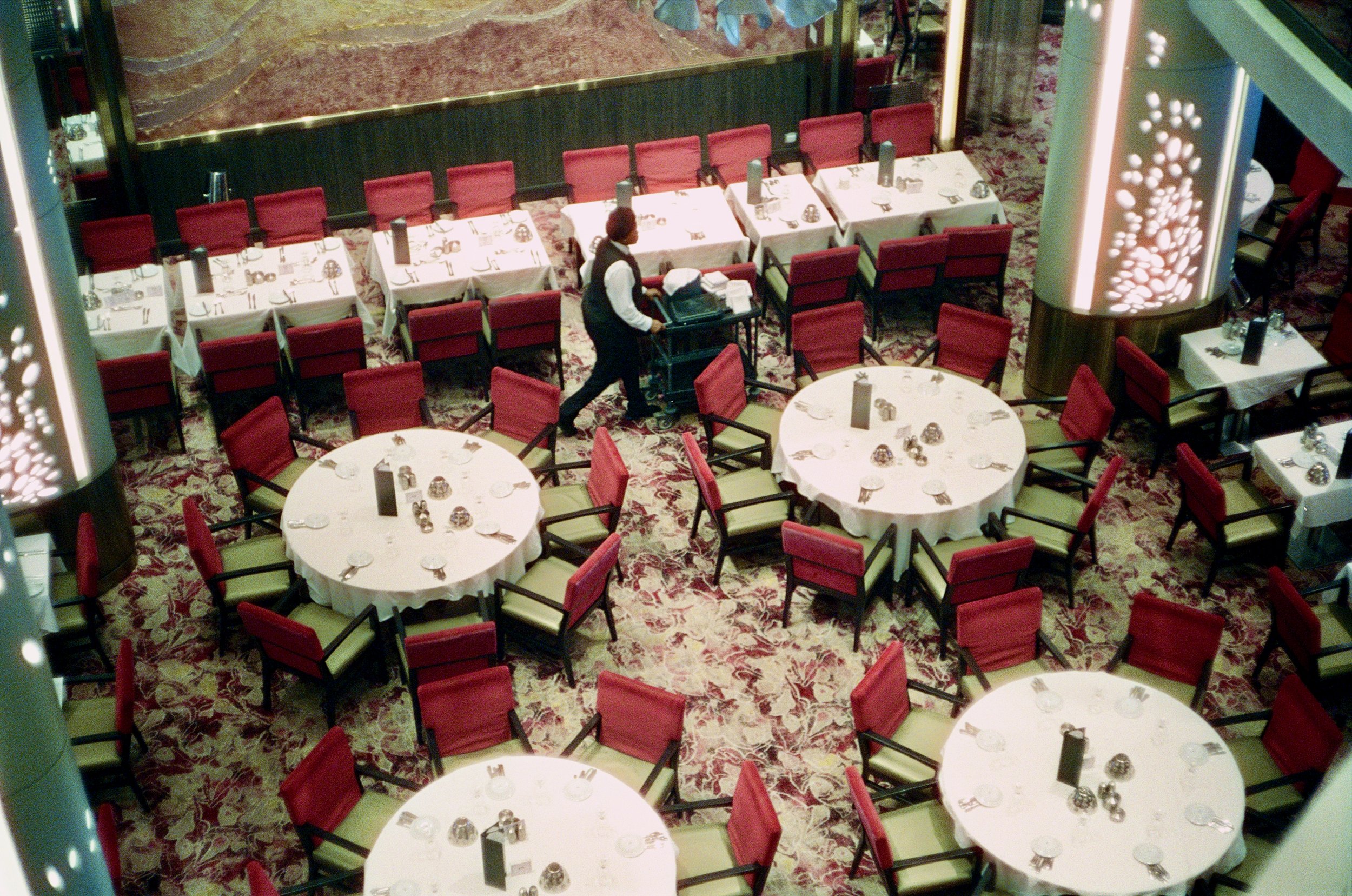 Empty restaurant dining area with round tables covered in white tablecloths, set with utensils and small glass centerpieces, surrounded by red chairs, and a waitress pushing a cart.