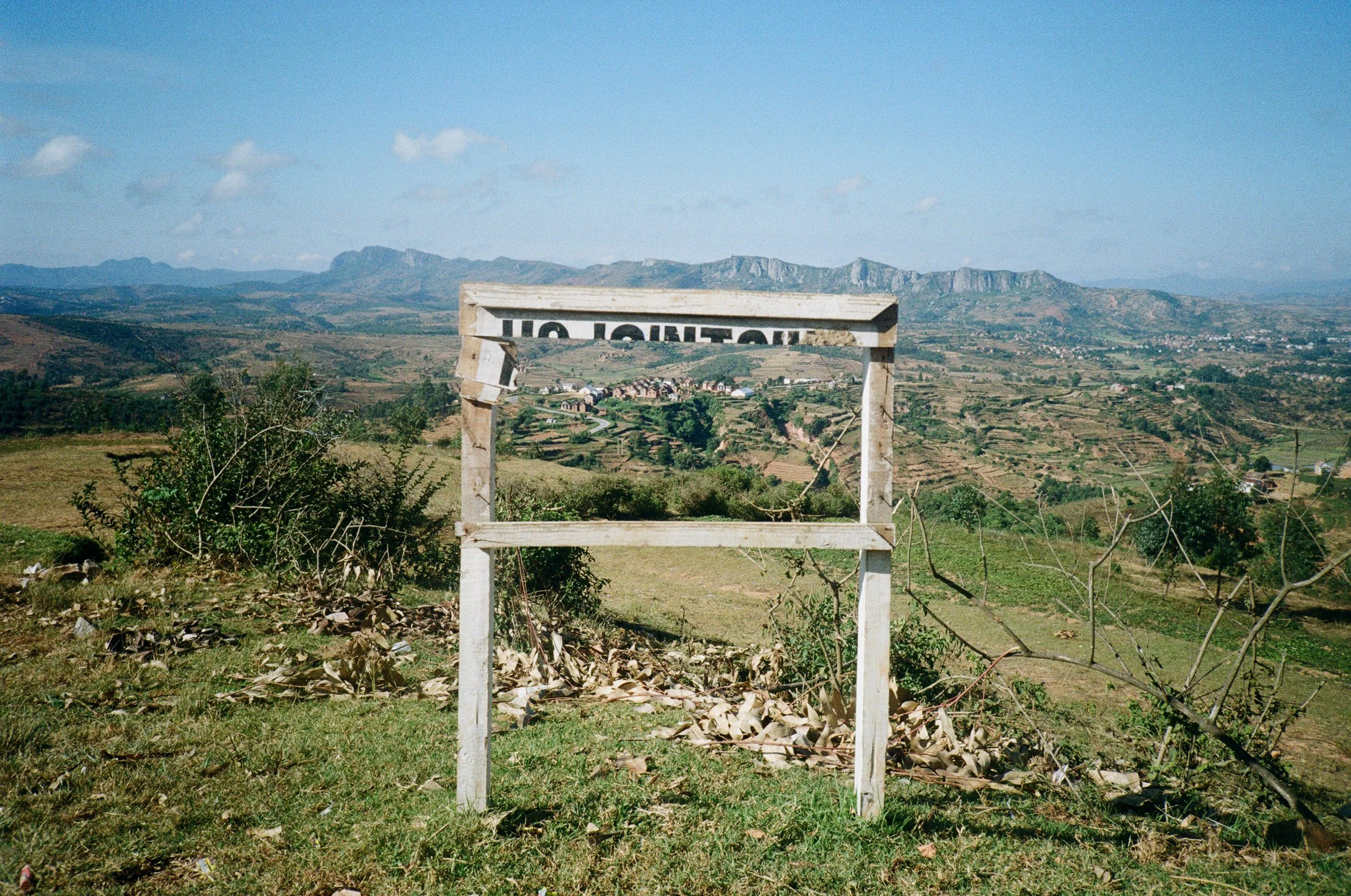 Scenic view of a rural landscape with a wooden frame in the foreground, rolling hills, small buildings, and mountains in the background under a blue sky with some clouds.