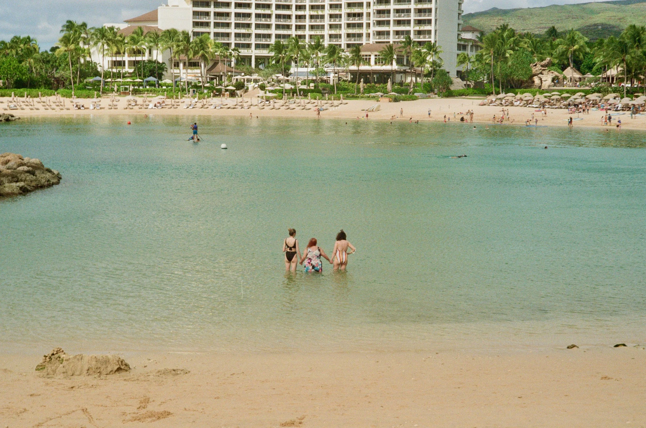 Three women in swimsuits walking into the water at a beach, with a hotel and green mountain in the background.
