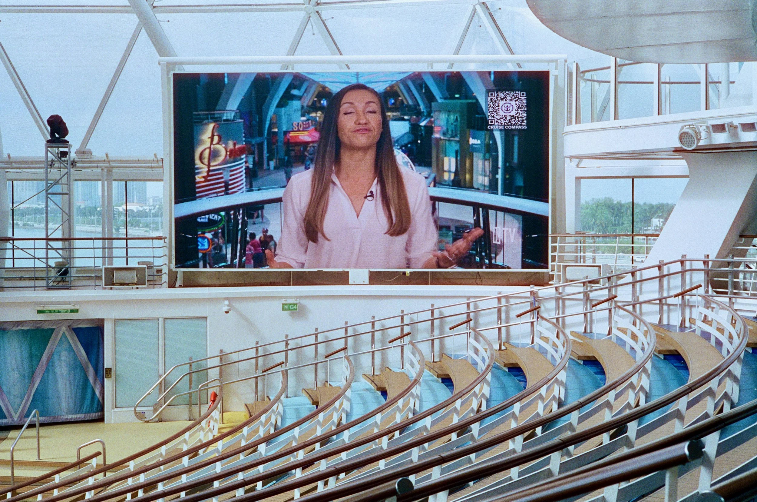 Large indoor area with curved seating and white railings, featuring a big digital screen displaying a woman speaking with an architectural background and a QR code, during daytime with natural light.