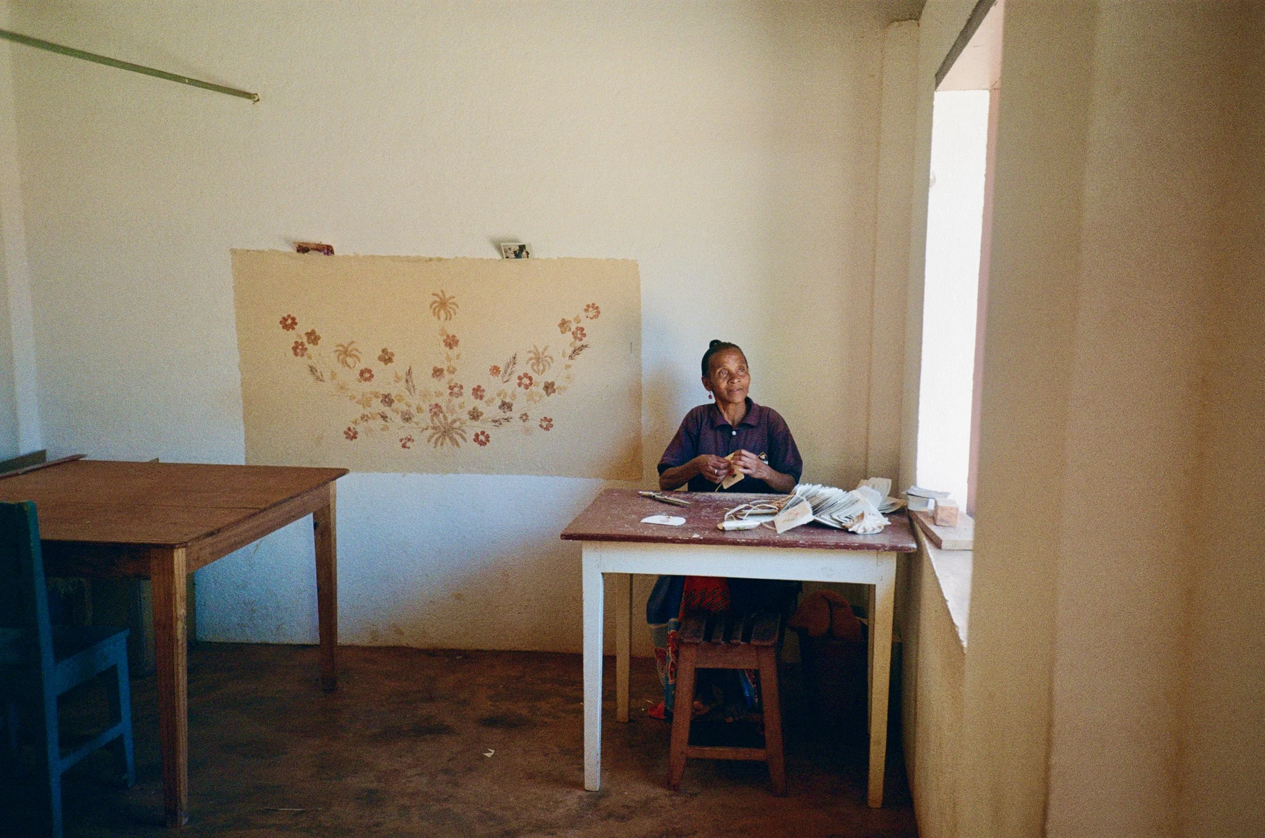 An elderly woman sitting at a desk in a sparsely decorated room, with a floral pattern on the wall behind her and wooden furniture around.