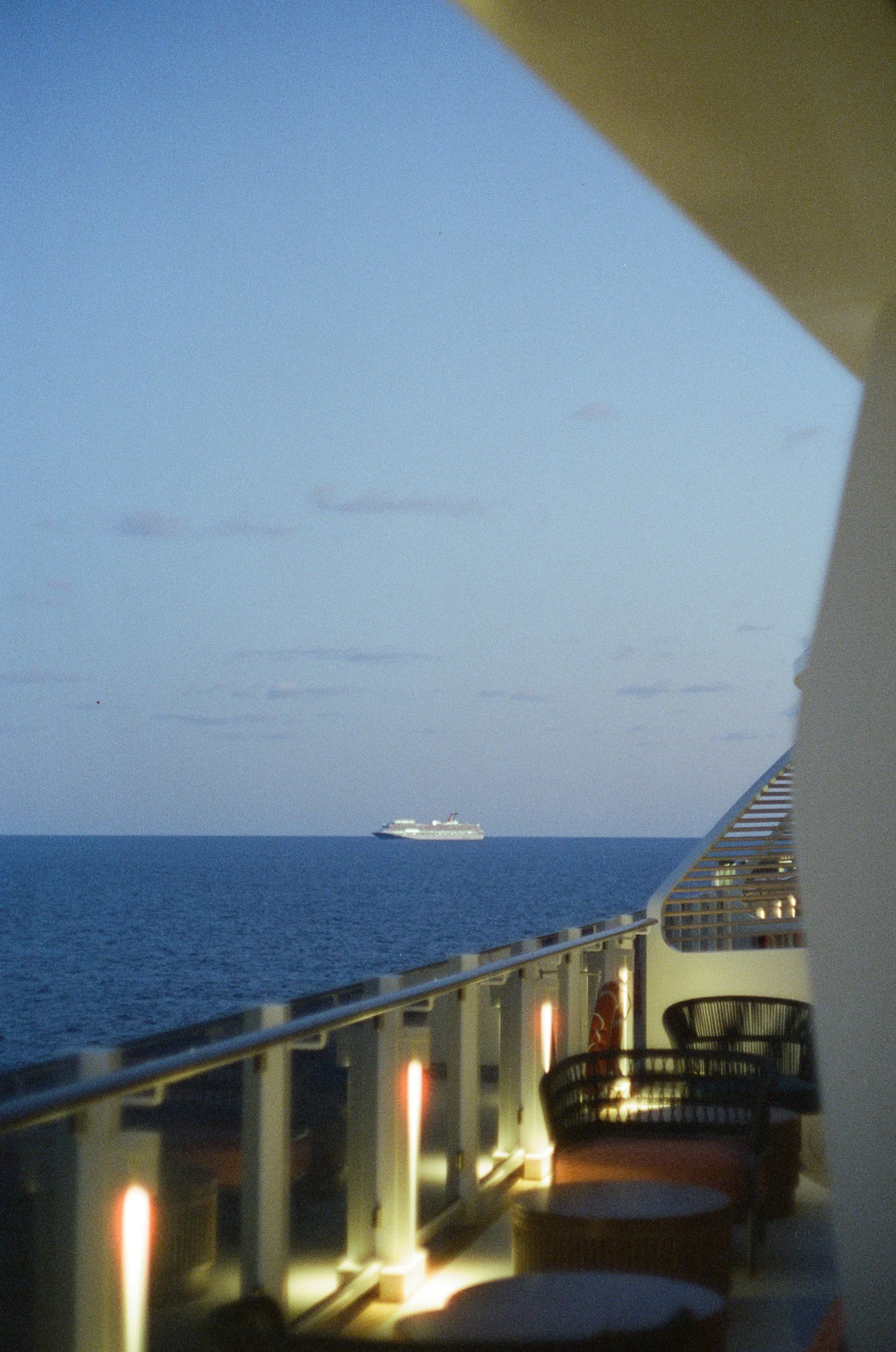 View from a cruise ship balcony showing the ocean, a cruise ship in the distance, and part of the ship's balcony and seating area at dusk.