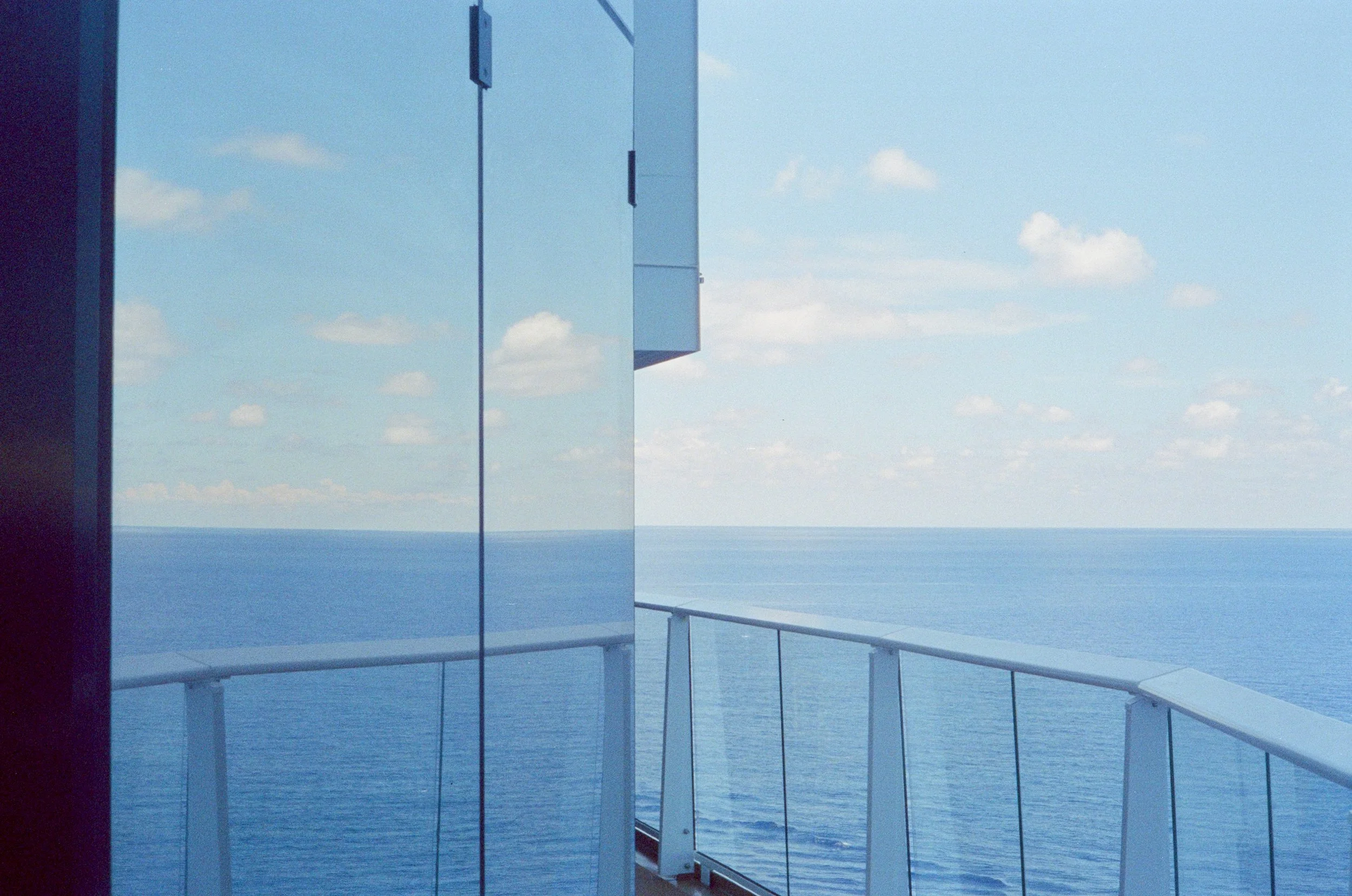View of the ocean from a balcony with reflective glass walls and a safety railing, under a partly cloudy sky.