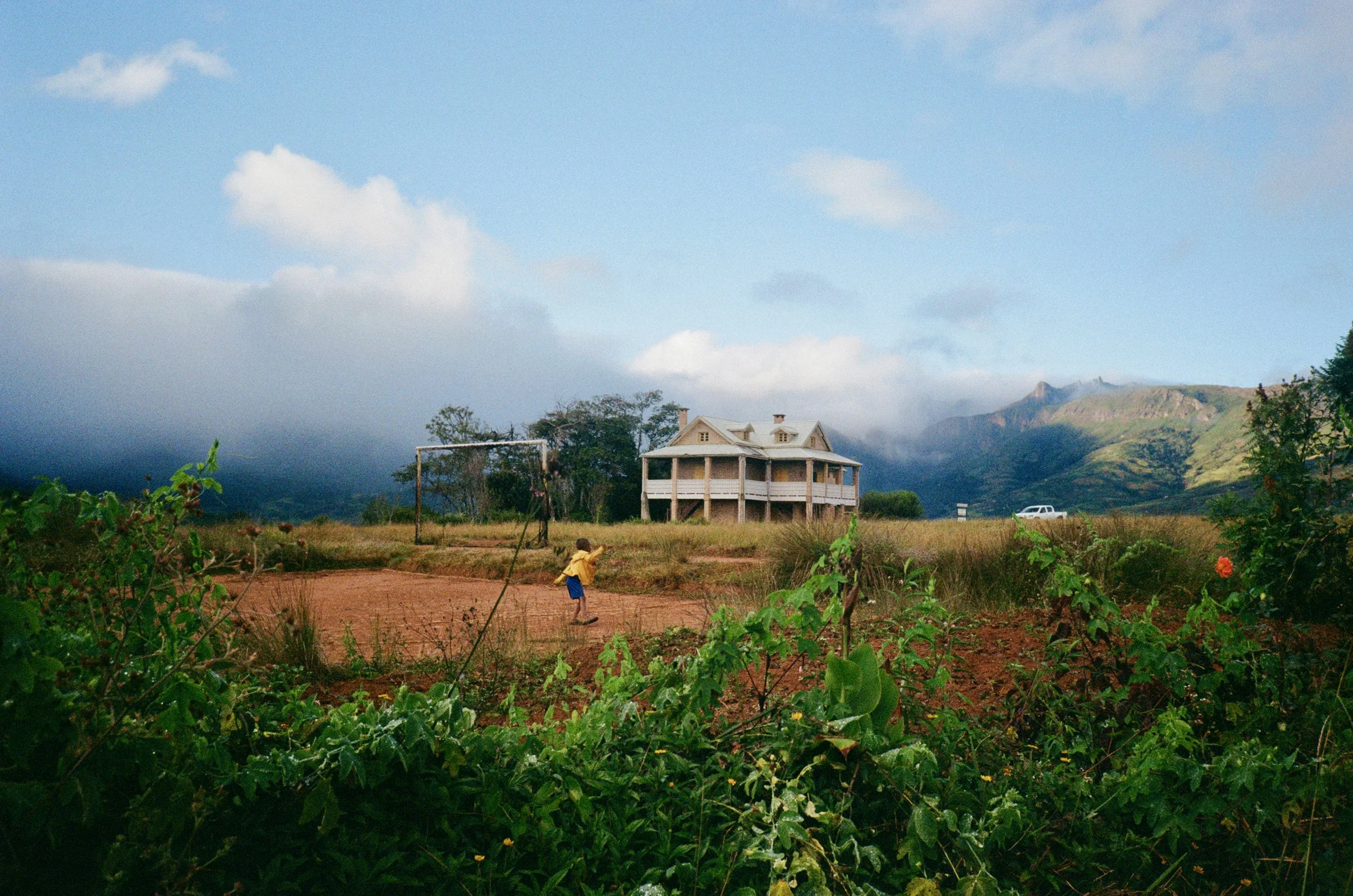 A rural landscape with a house on stilts in the background, green vegetation in the foreground, a young child playing on the dirt ground, and cloudy sky with some mountains in the distance.