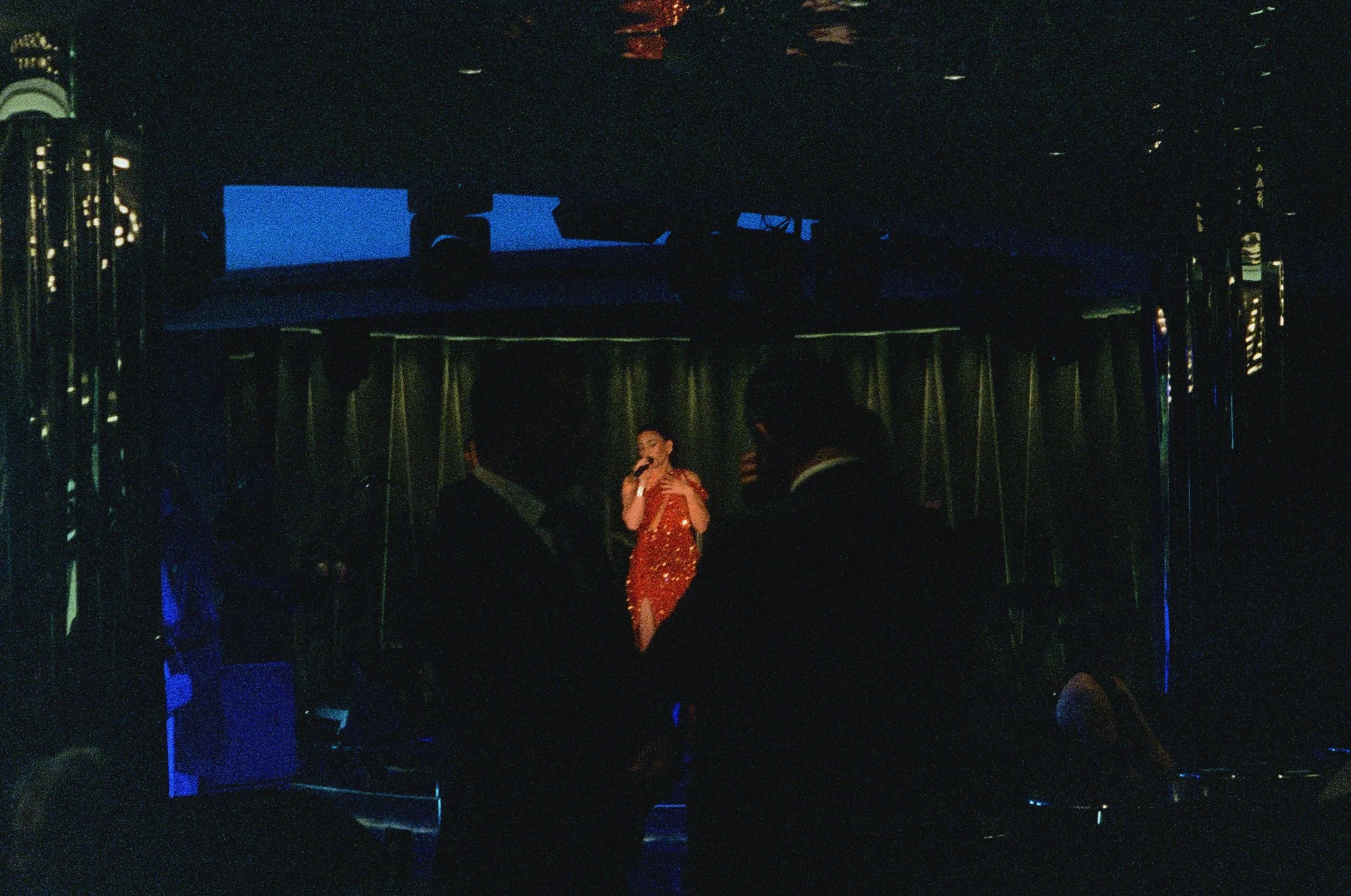 A singer in a red glittery dress performing on stage with a microphone in hand, surrounded by dim lighting, curtains, and reflective vertical decor.