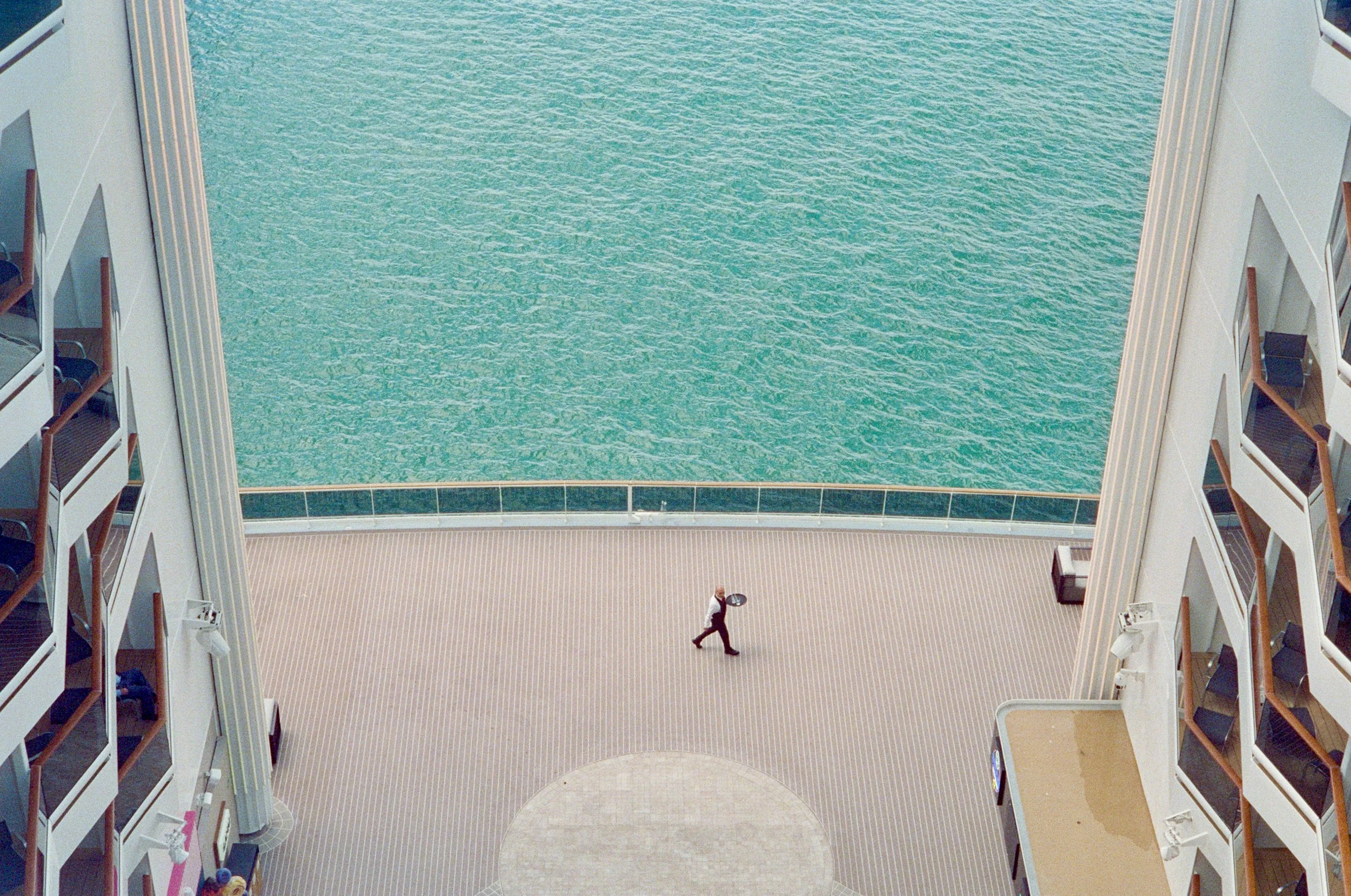 View from inside a high-rise building, showing an atrium with a large pool of water at the top floor, surrounded by balconies and doorways on each side, and a person walking on the lower level.