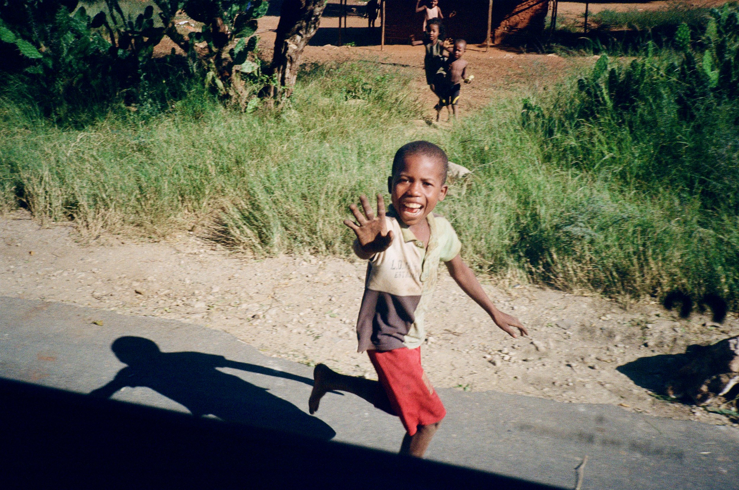 Two boys running on a dirt road, one in a light-colored shirt and red shorts waving, with another boy and a girl in the background near a fence and a tree, surrounded by green vegetation.