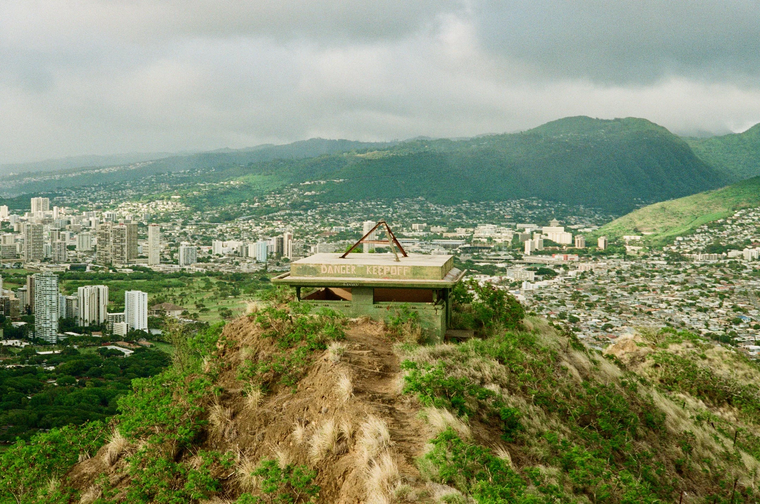 View of a cityscape with tall buildings, green hills, and mountains in the background under cloudy skies; a wooden overlook with a 'Danger Keep Off' sign in the foreground.