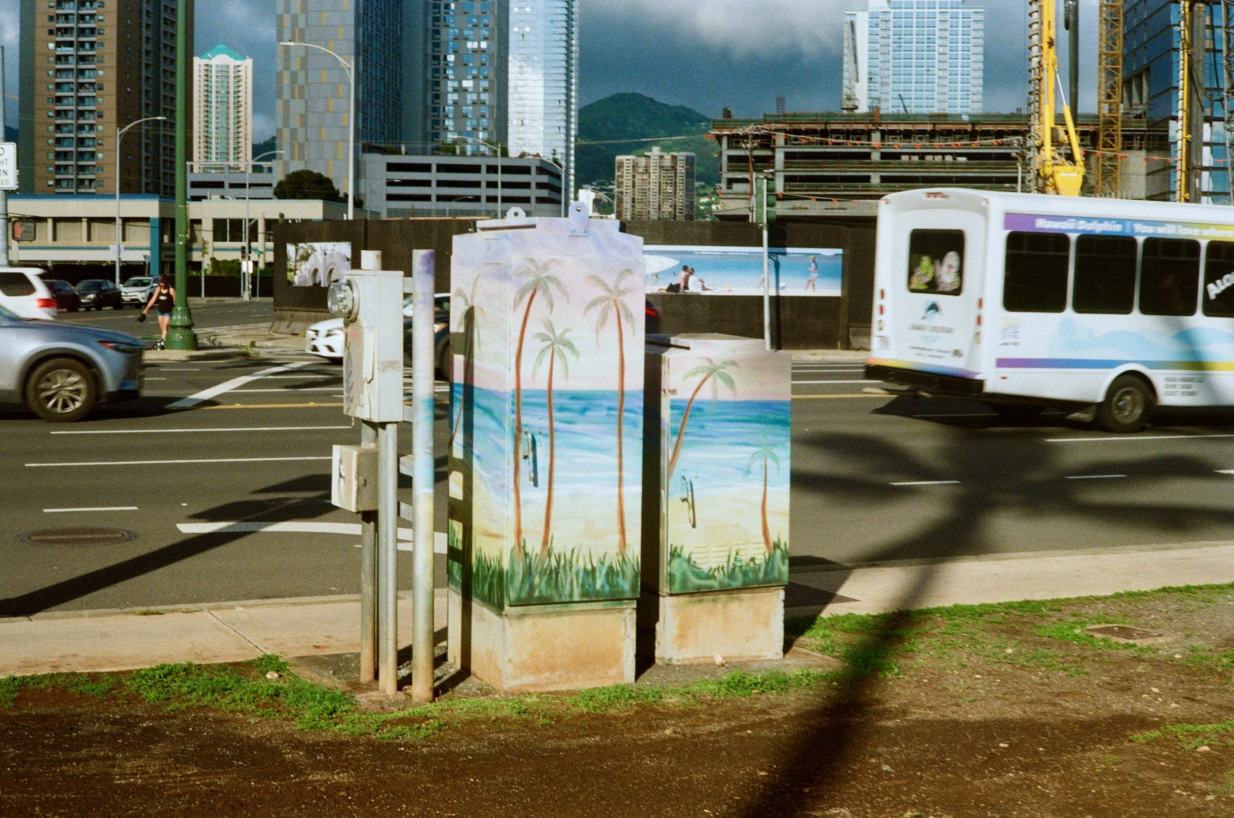 City street scene with tall modern skyscrapers, construction site, moving trolley, cars, person walking on sidewalk, and colorful painted utility box with beach palm trees