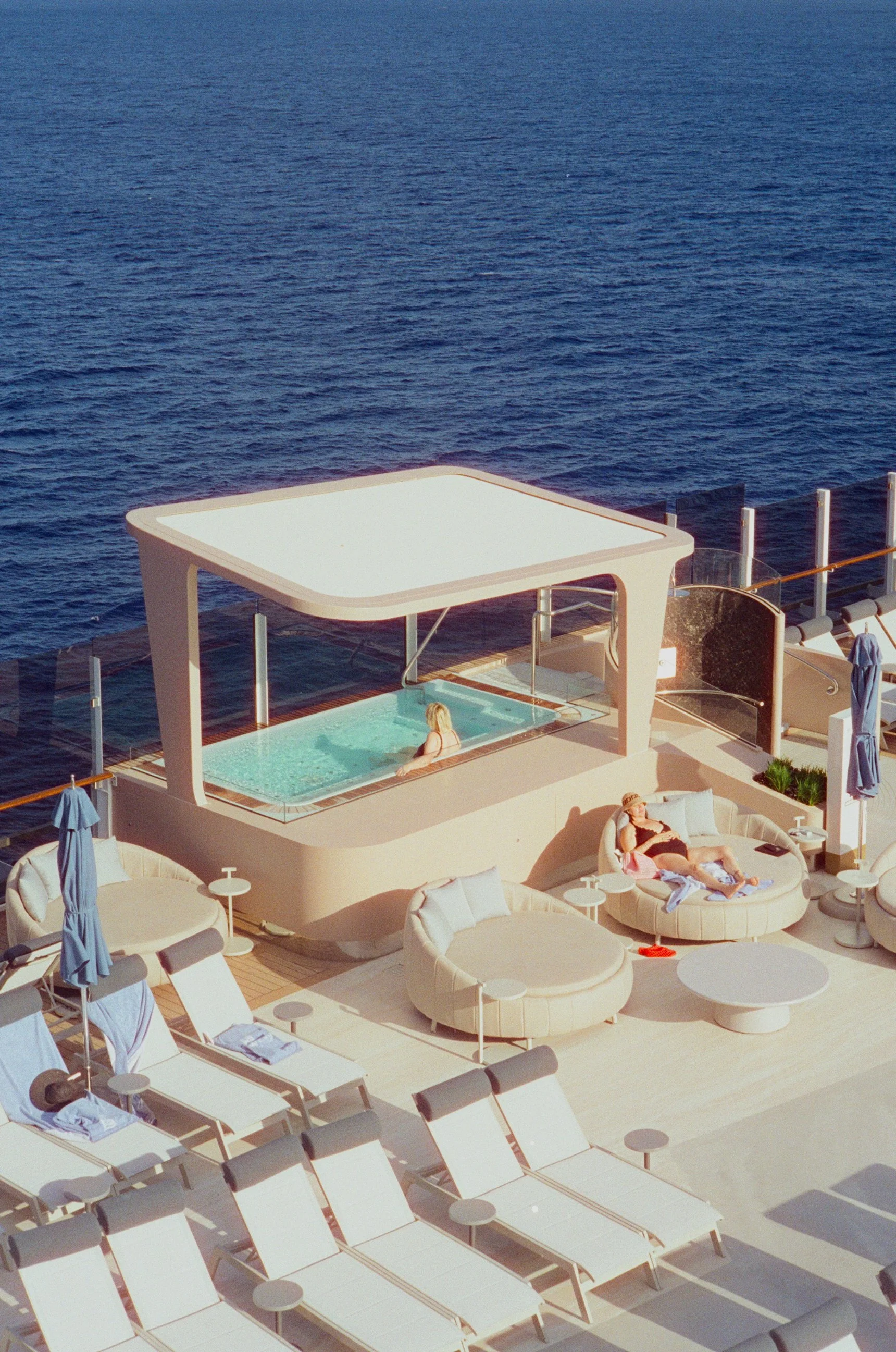 A woman relaxing in a small pool on a cruise ship deck, with ocean water in the background and surrounding lounge chairs and umbrellas.