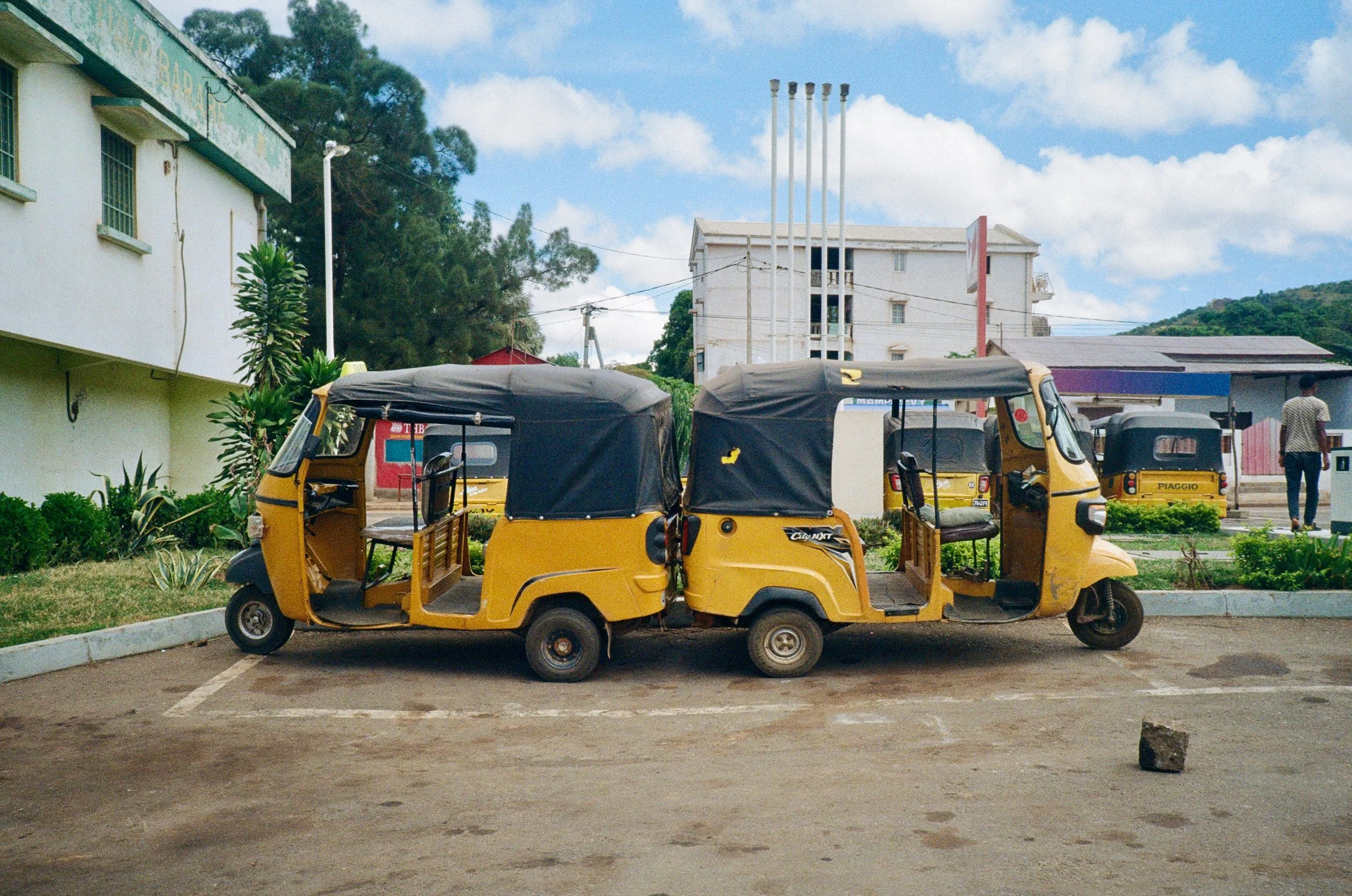A yellow auto rickshaw parked in a parking lot with a building and trees in the background under a partly cloudy sky.