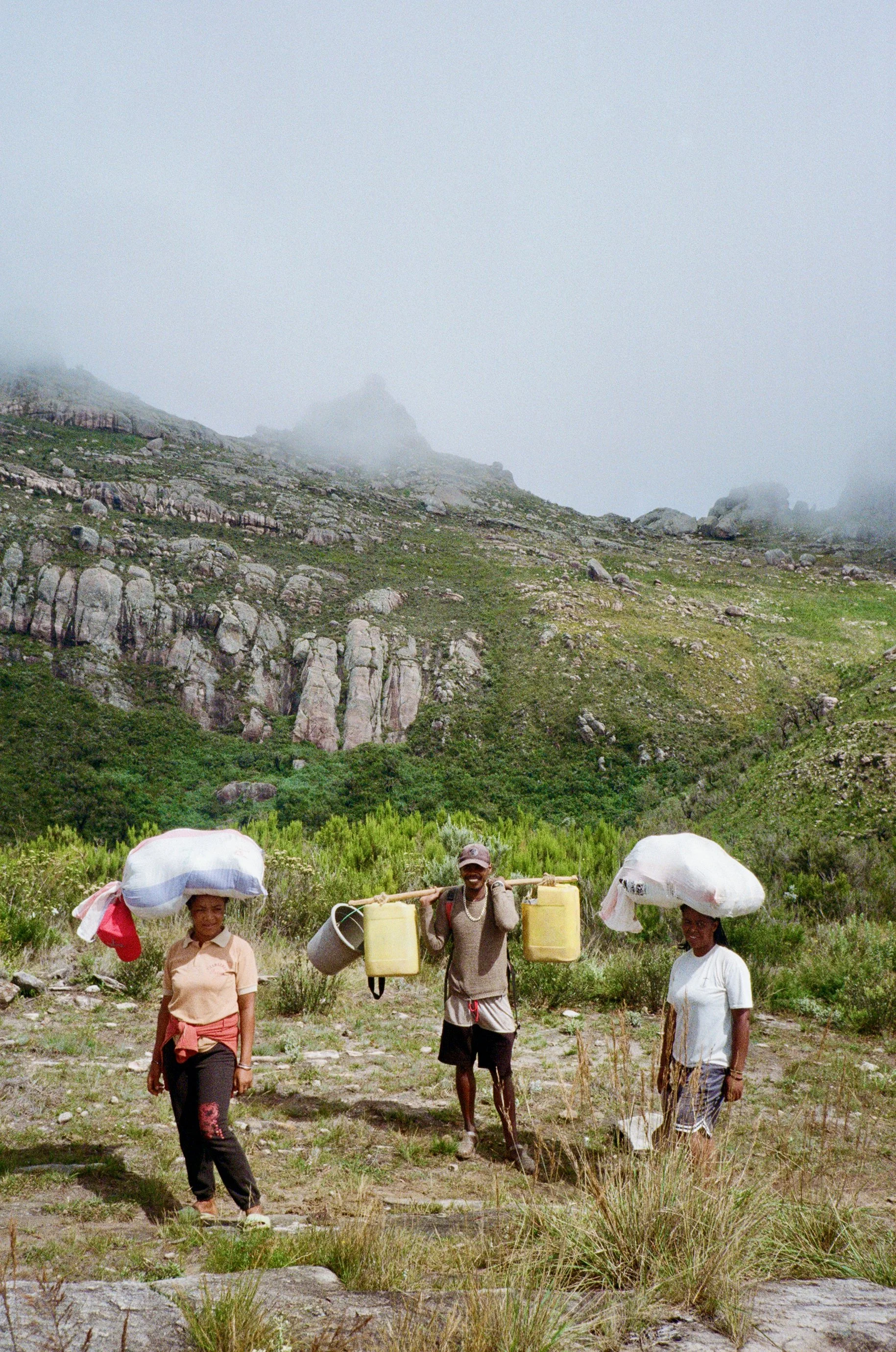Three people, two women and one man, are walking in a grassy area with mountains and fog in the background. They carry umbrellas and containers, and appear to be working or traveling in a rural outdoor setting.