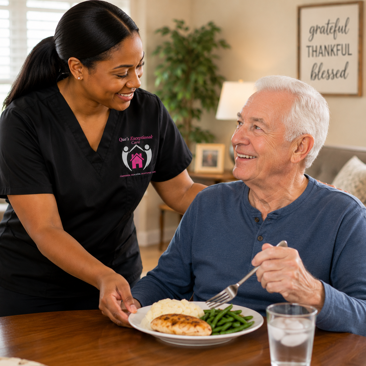 A smiling home caregiver in black scrubs assists an elderly man with his meal at a dining table. The man has white hair, wears a blue long-sleeve shirt, and is holding a fork. The meal includes green beans, mashed potatoes, and a piece of cooked chicken. In the background, there is a framed quote that says 'grateful, thankful, blessed,' and a cozy living room setting.