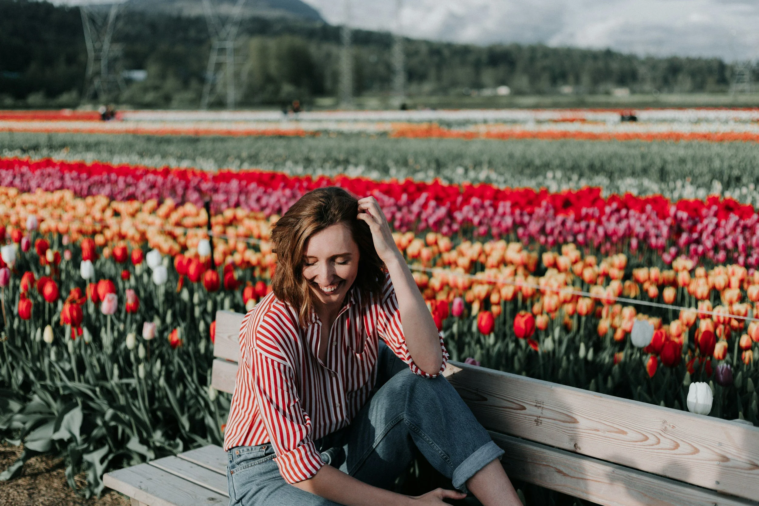 A woman with brown hair wearing a red and white striped shirt sitting on a wooden bench in a tulip field with colorful tulips and mountains in the background.