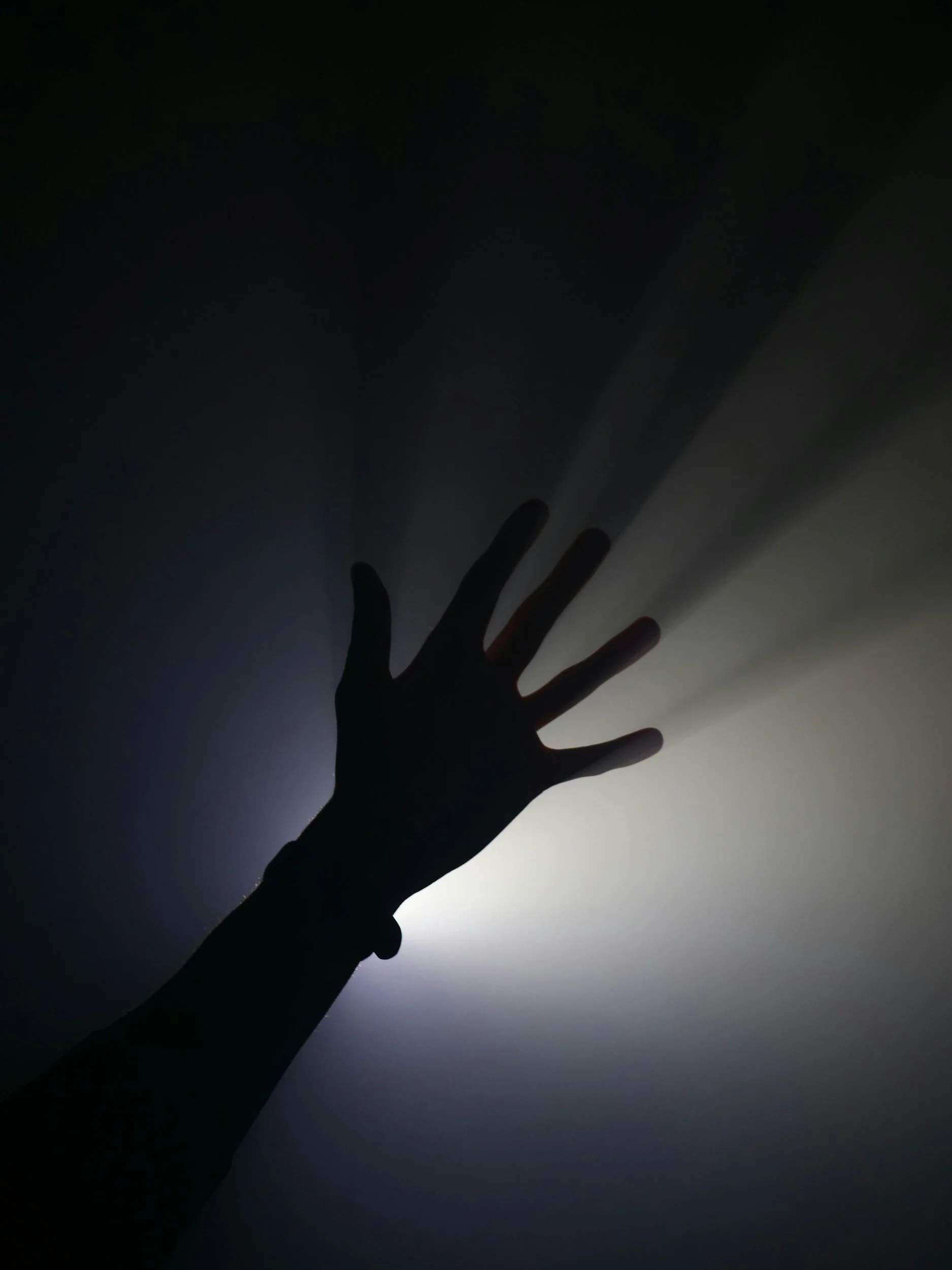 Silhouette of a hand reaching toward the sky with a background of dark fog and light source creating a glow.