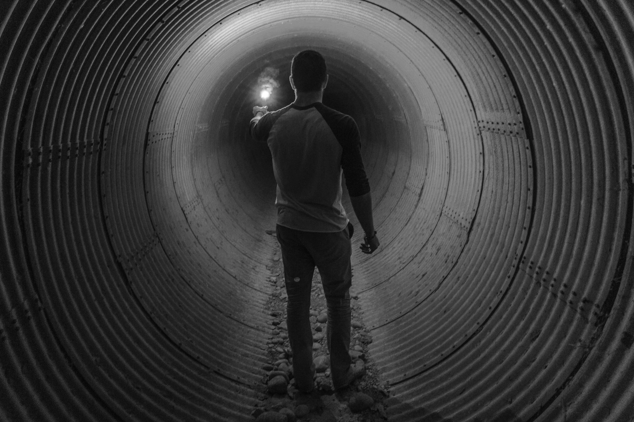 A person walking through a large metal pipe tunnel, holding a flashlight in the distance, with gravelly ground underneath.