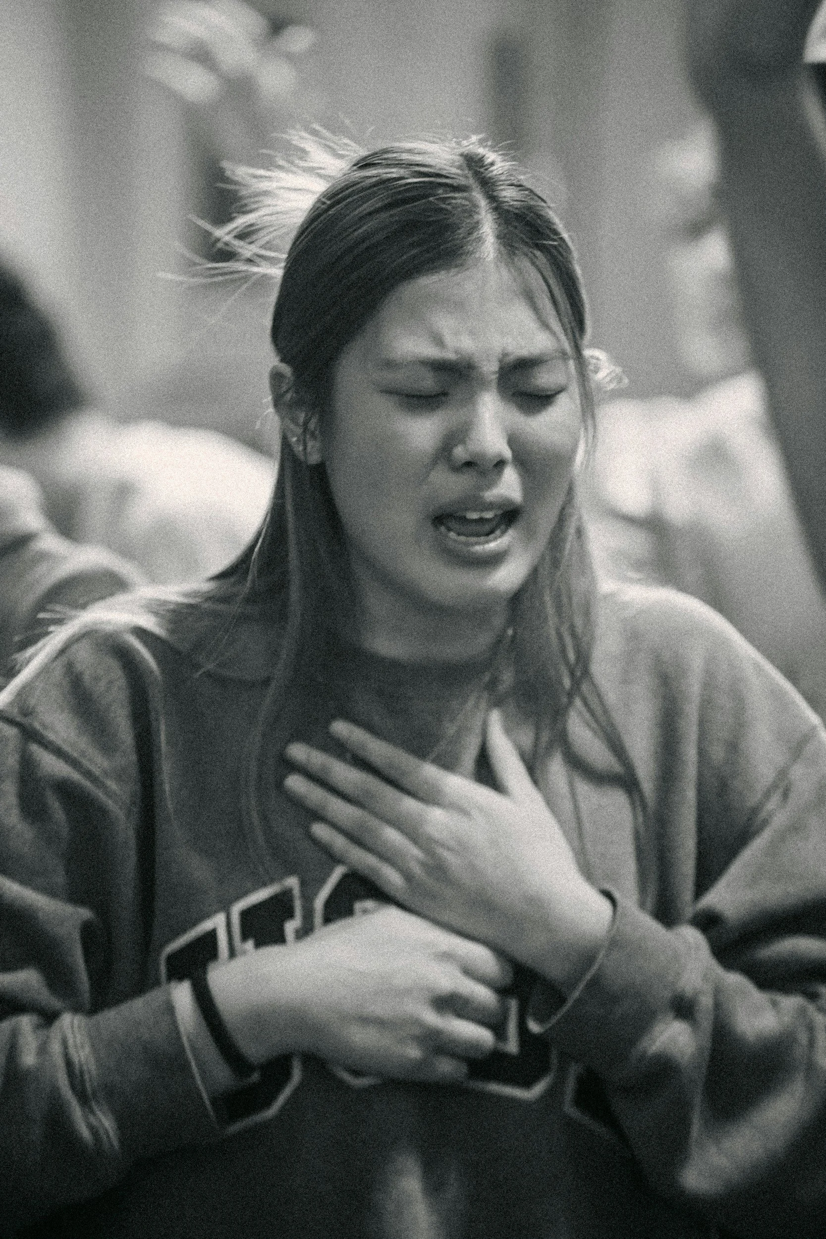 A black and white photo of a young woman with her eyes closed, hand over her chest, appearing emotional or distressed, wearing a sports jersey.