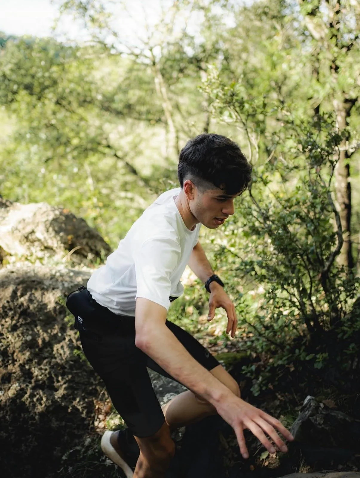 Joven en camisa blanca y pantalones cortos negros recolectando o inspeccionando algo en el bosque. El fondo tiene árboles y vegetación.