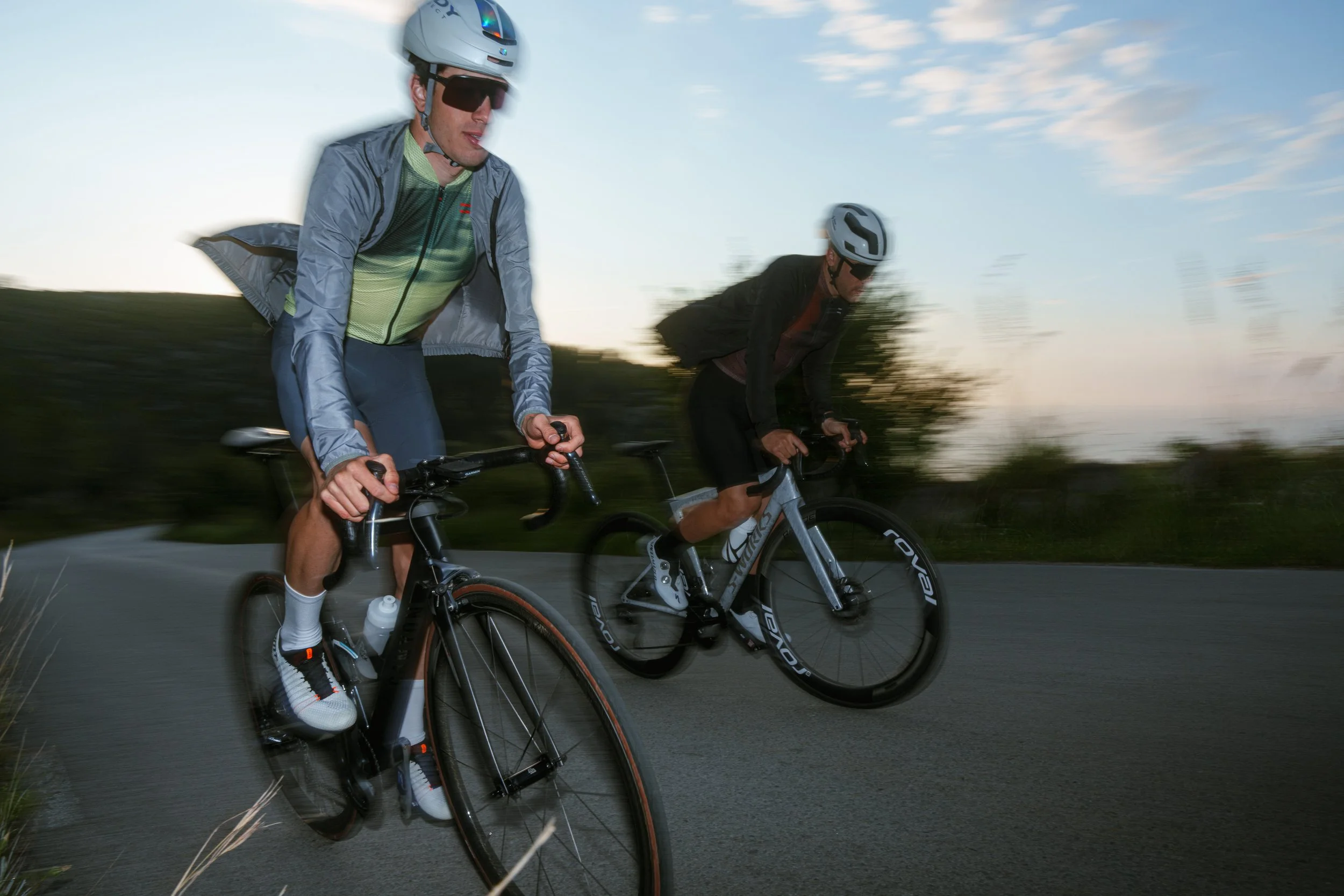 Dos ciclistas pedaleando en una carretera con fondo de cielo y nubes, uno lleva casco con visor oscuro y otro con casco blanco y gafas, ambos con ropa deportiva y accesorios para ciclismo.