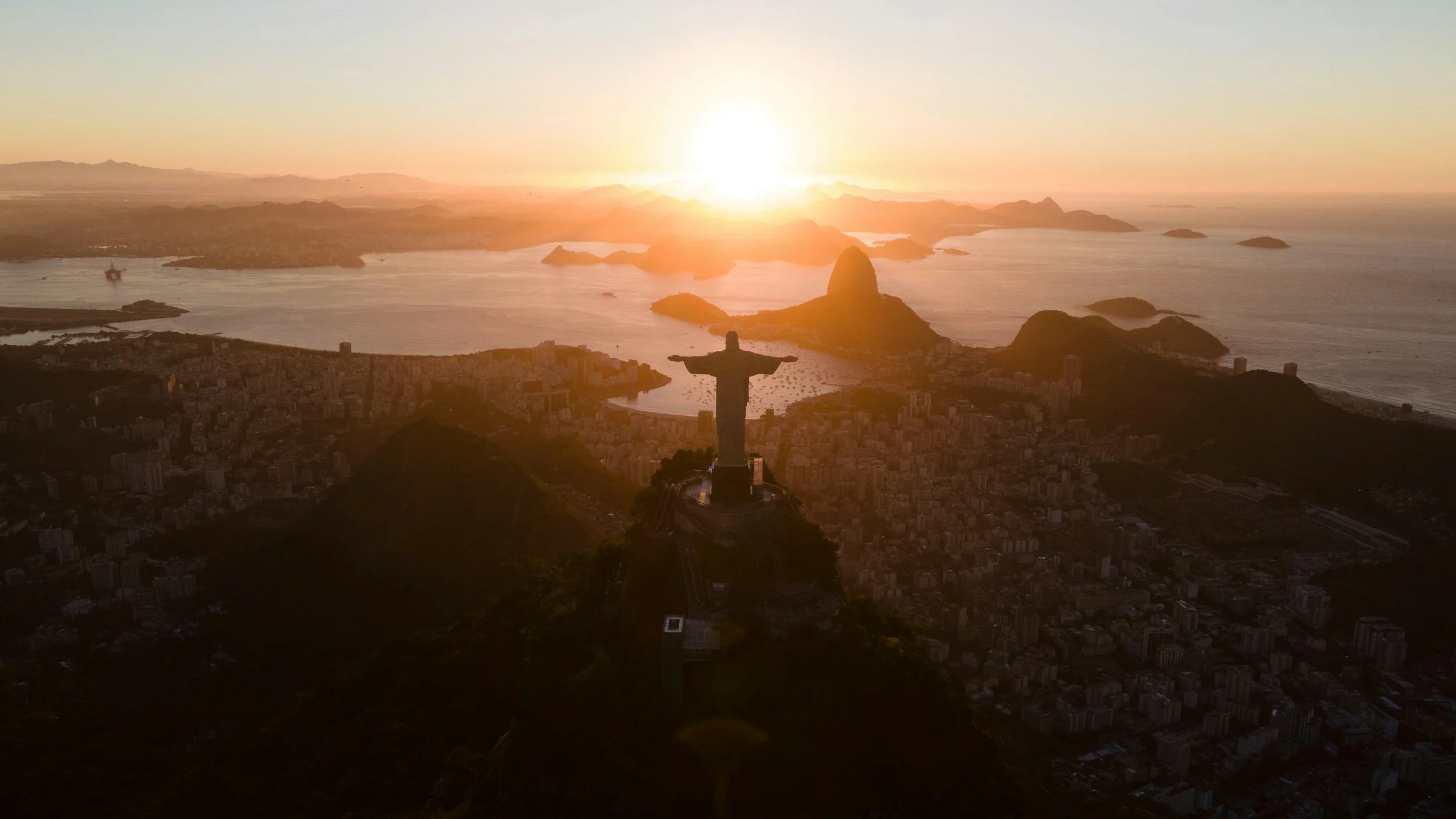 View of Rio de Janeiro at sunset featuring the Christ the Redeemer statue in the foreground and Sugarloaf Mountain in the background.