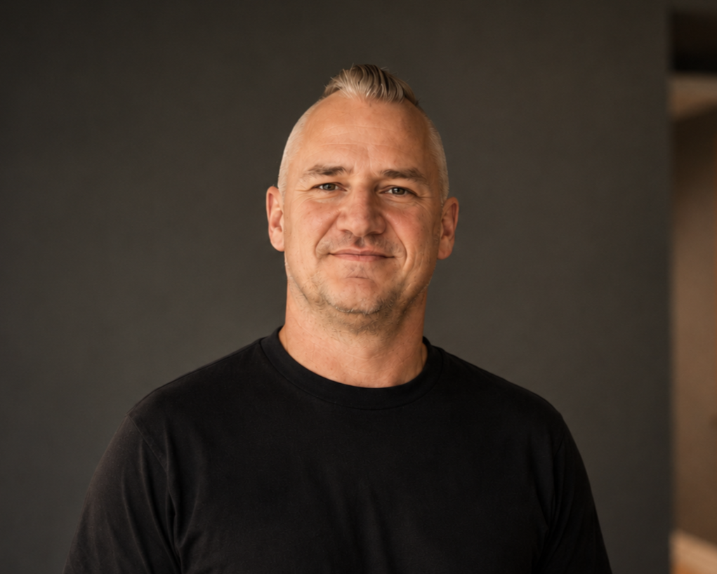 A man with a short, styled blonde haircut wearing a black t-shirt standing in front of a plain dark background.