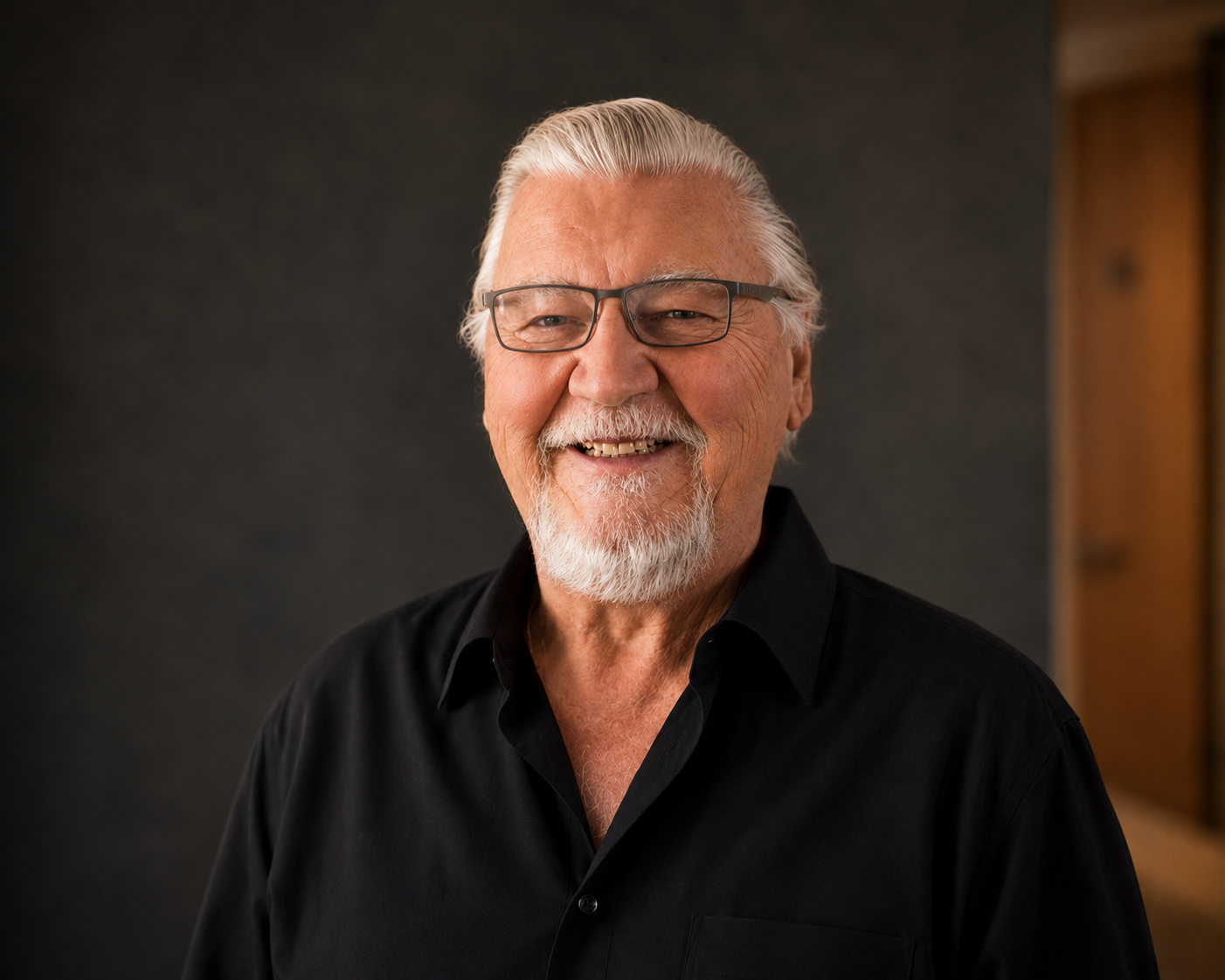 Smiling elderly man with white hair, beard, and glasses, wearing a black shirt, standing indoors with a dark background and wooden panel on the right.