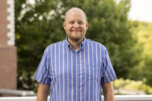 A smiling man with a beard wearing a blue, yellow, and white striped shirt, standing outdoors with green trees in the background.
