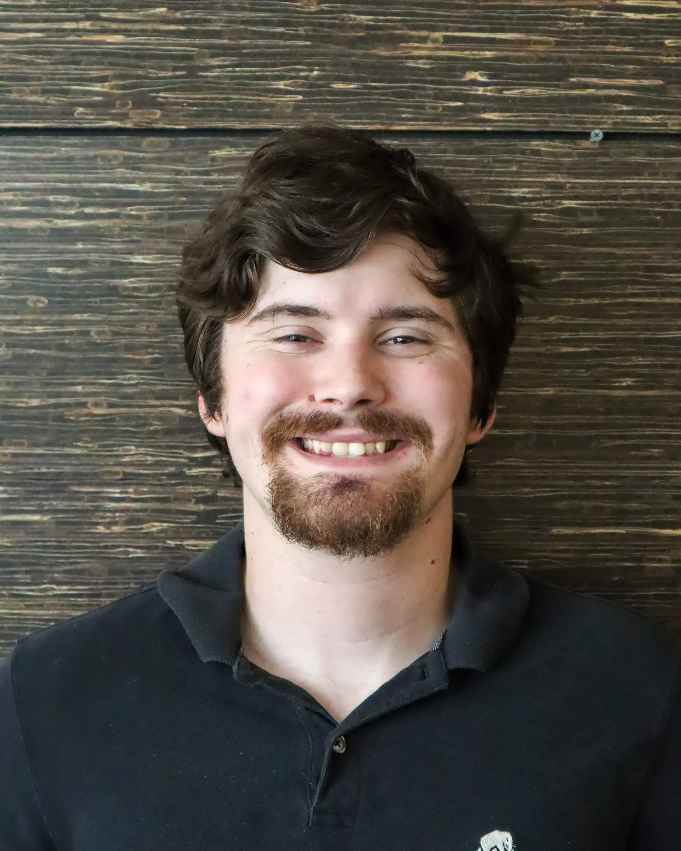 Smiling young man with dark wavy hair, facial hair, wearing a black hoodie, against a wooden wall background.