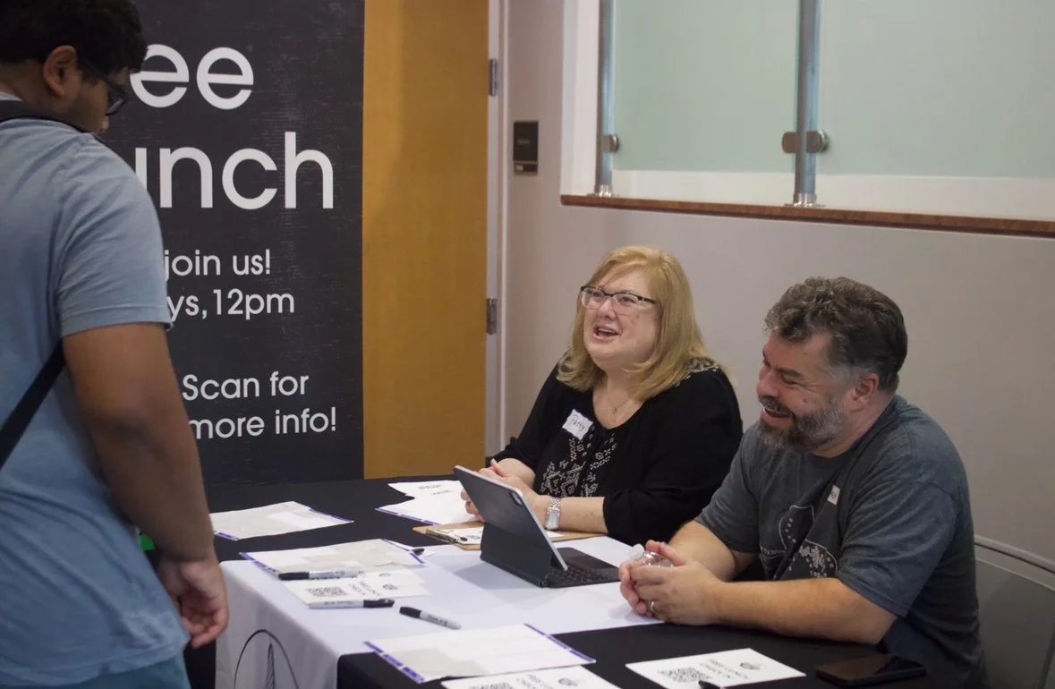 A man and a woman sitting at a table behind a sign, talking to a person standing in front of them. The table has papers, markers, and a tablet. The sign behind them promotes a lunch event at 12pm.