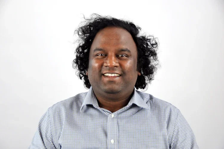 A man with curly black hair wearing a checkered button-up shirt, smiling at the camera, against a plain light-colored background.