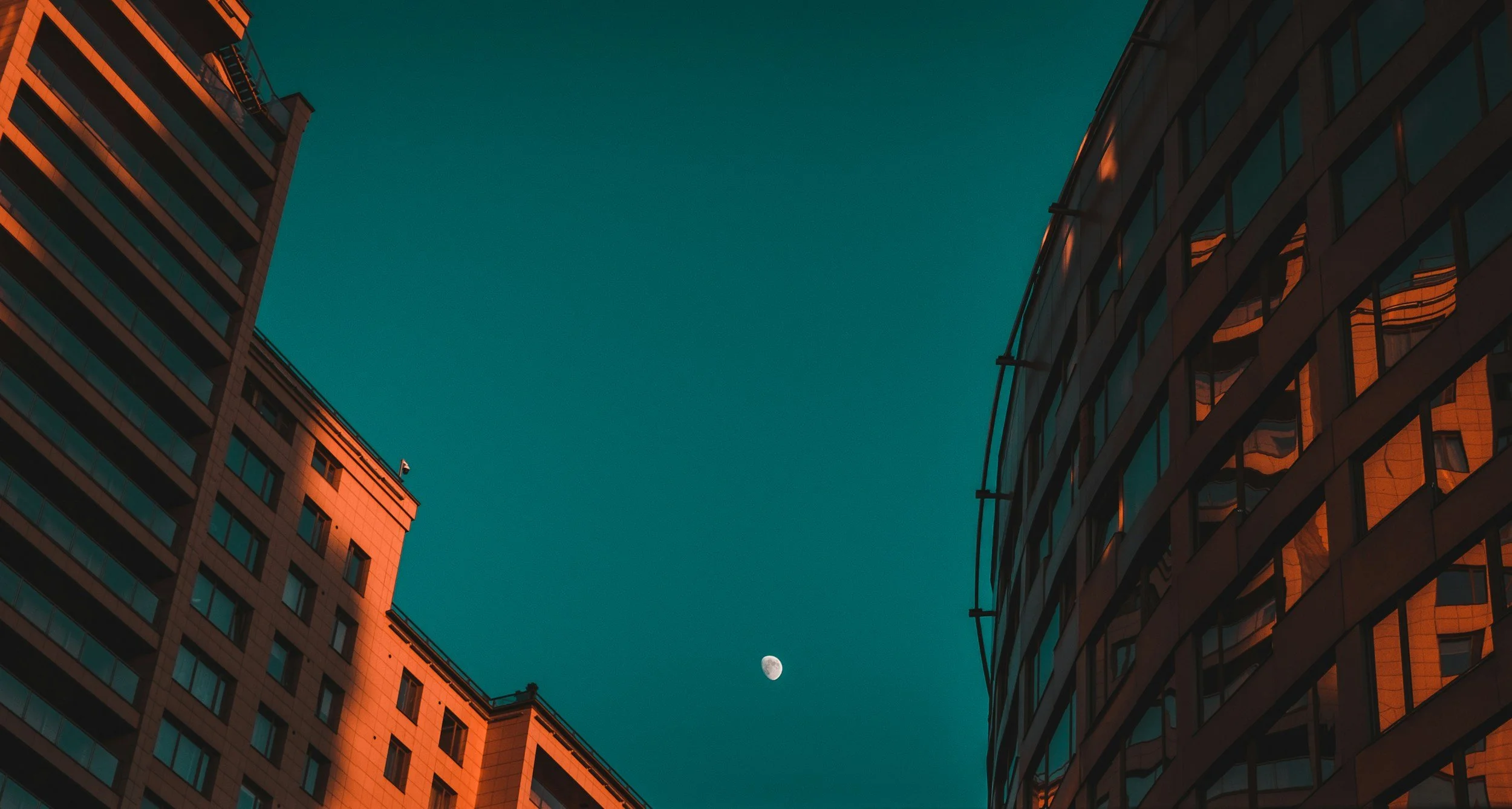View of two tall modern buildings with reflective glass windows at dusk, with the moon visible in the sky between the buildings.
