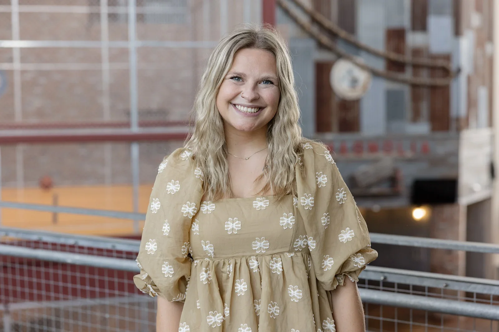 A smiling woman with blonde wavy hair, wearing a beige dress with white floral embroidery, standing outdoors on a balcony with a brick building and a roller coaster in the background.