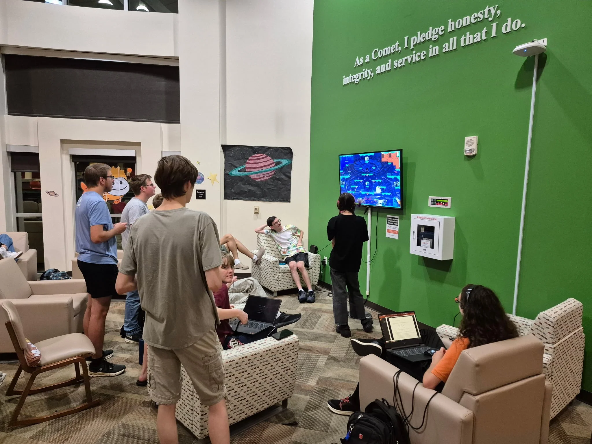A group of people waiting in a lounge area at an indoor event space, with some standing and others sitting on chairs or couches. There is a bright green wall with white text on it, and a television screen displaying a game or digital content. The lounge is decorated with space-themed posters, and there are laptops on people's laps.