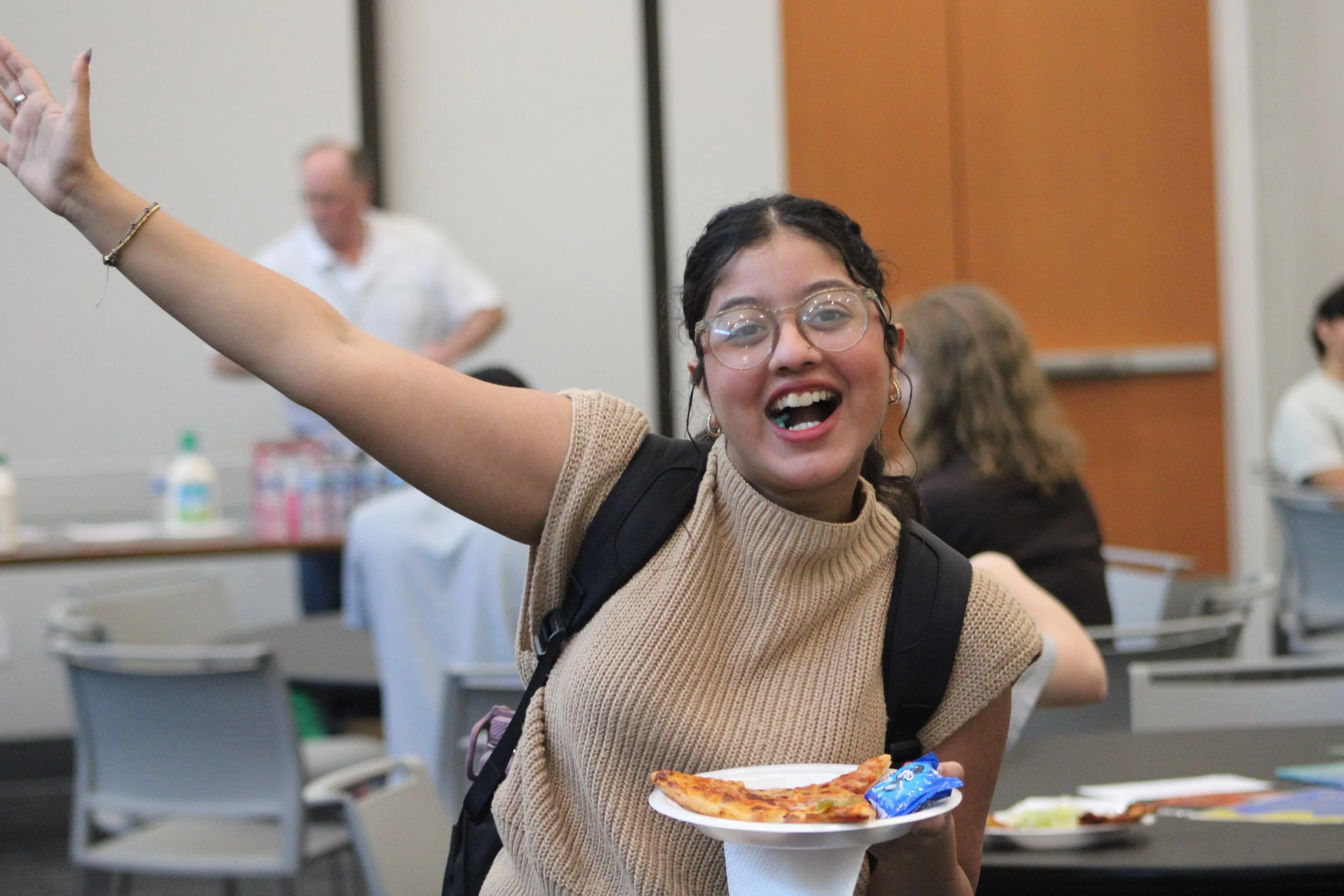 A young woman with glasses and dark hair, smiling and raising her arm, is holding a plate with a slice of pizza and a snack in a classroom or conference setting.
