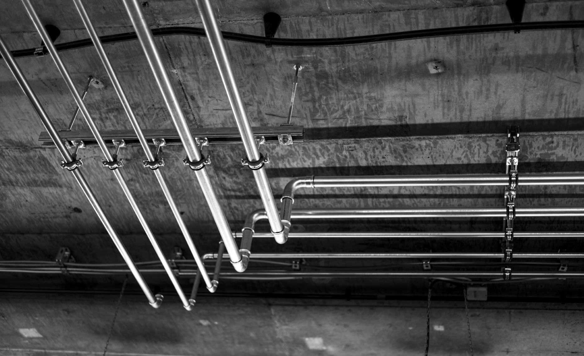 Black and white photo of metal pipes mounted on a concrete ceiling, with some pipes bent at right angles.
