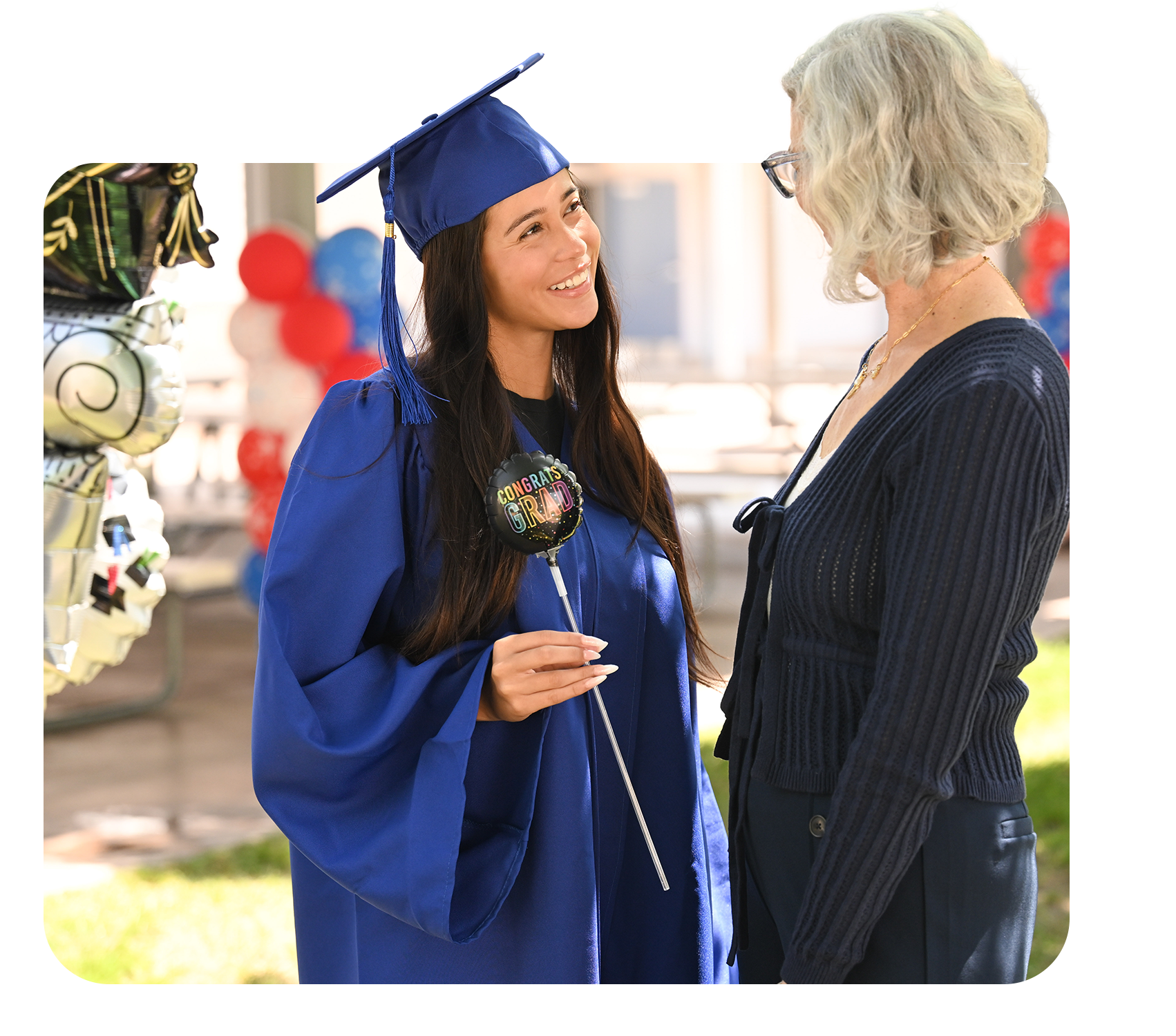 A young woman in a blue graduation gown and cap smiling and holding a black balloon that says "Congrats Grad" while talking with an older woman wearing glasses and a dark blue cardigan, outdoors at a graduation celebration with balloons in the background.