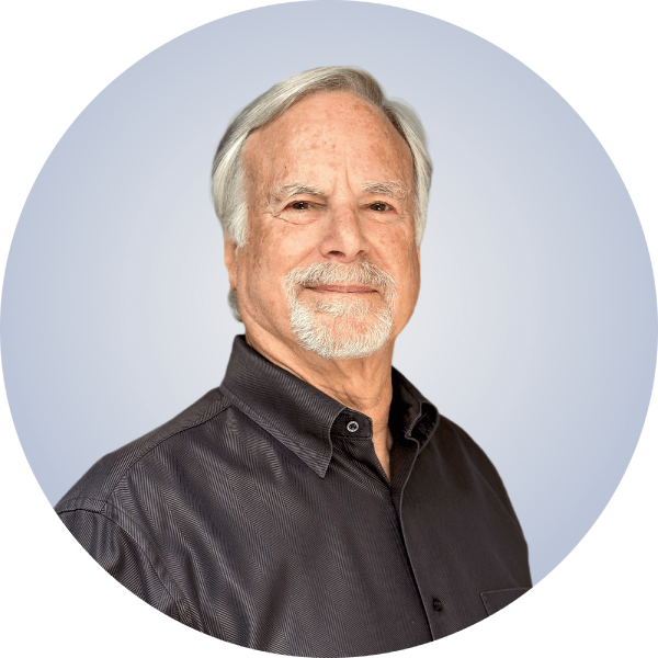 An older man with gray hair and a beard, wearing a black button-down shirt, smiling at the camera against a light blue background.