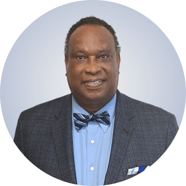 A man with dark skin and short curly hair, wearing a dark checked blazer, a light blue shirt, and a matching blue bow tie, standing against a plain light background.