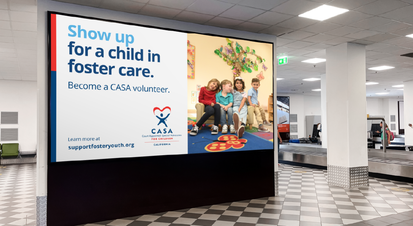 Large digital billboard in an airport with a message encouraging people to become CASA volunteers for foster children. The ad features a photo of four children sitting on a bench in a classroom, smiling and looking happy.